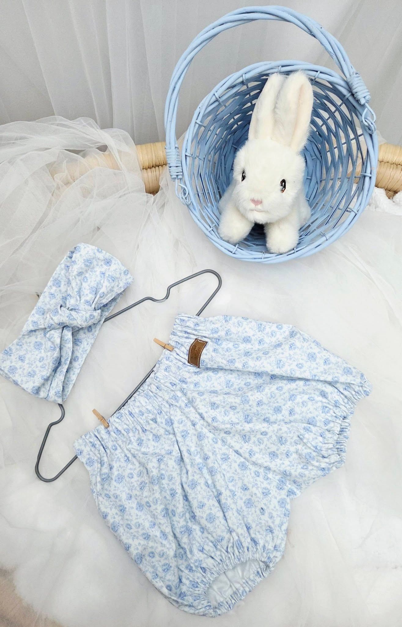 Blue floral baby romper with matching headband on hanger, white bunny plush toy in blue wicker basket.