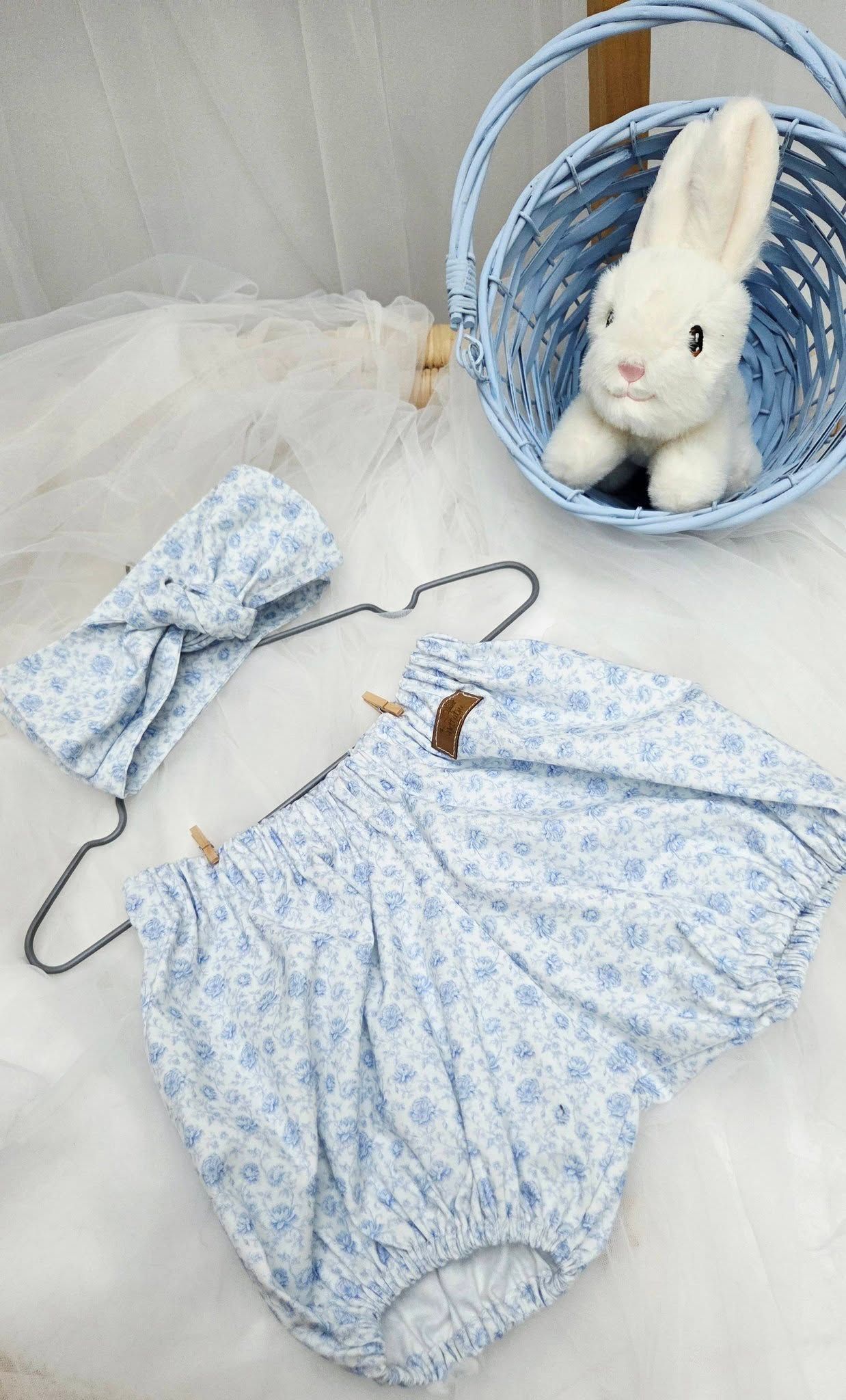 Blue floral baby bloomers and headband set on hanger beside a white stuffed bunny in a light blue wicker basket.