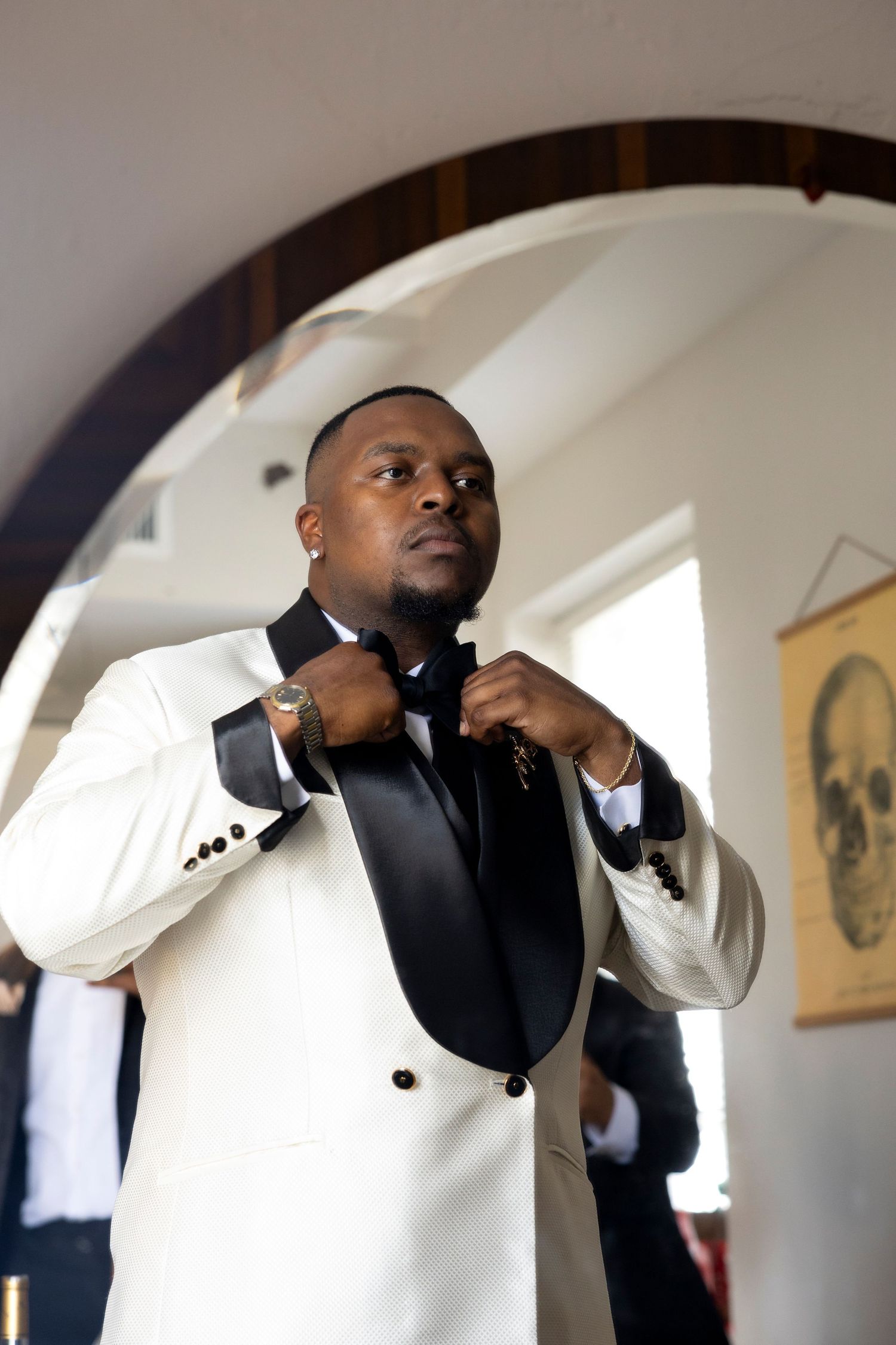 A person adjusts their cream and black tuxedo jacket while getting dressed in an elegant setting with arched doorways.