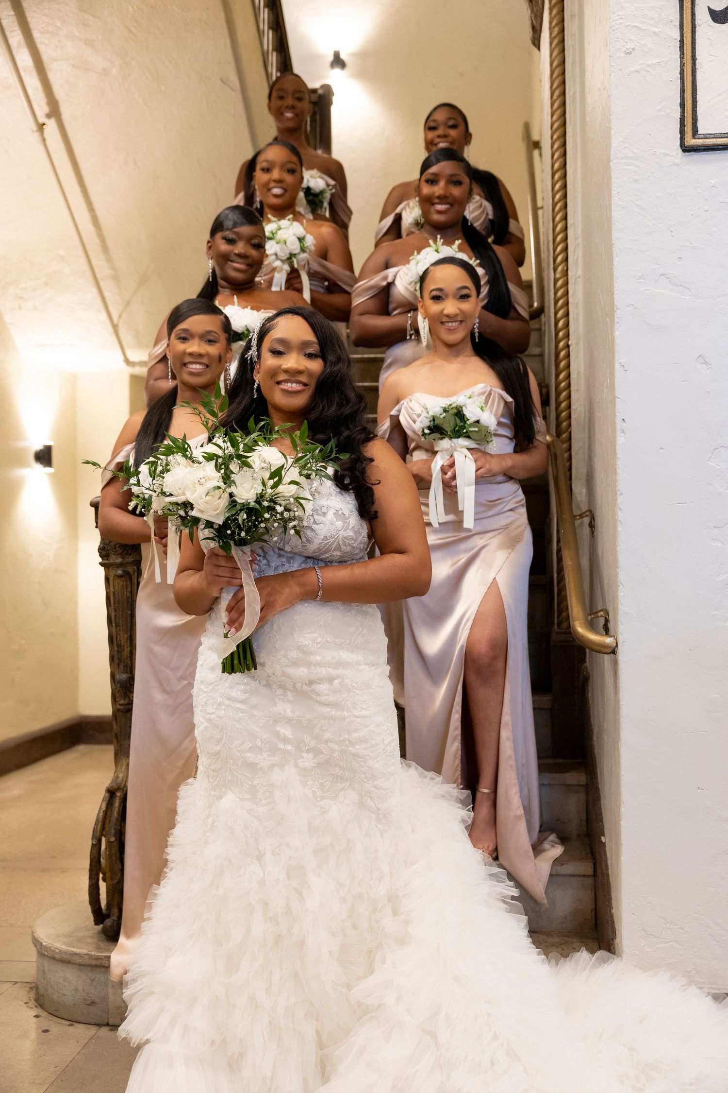 Bridal party poses joyfully on elegant staircase with bridesmaids in blush pink dresses holding white bouquets.