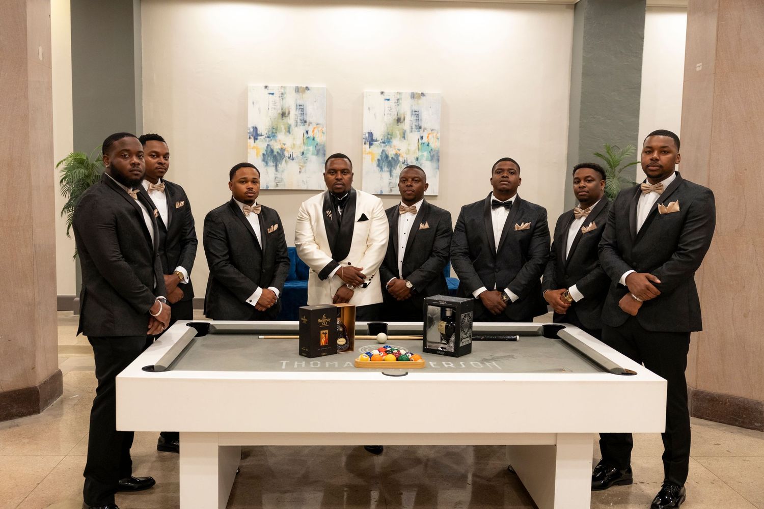 Groomsmen in black tuxedos gather around a white table at an elegant indoor reception venue.