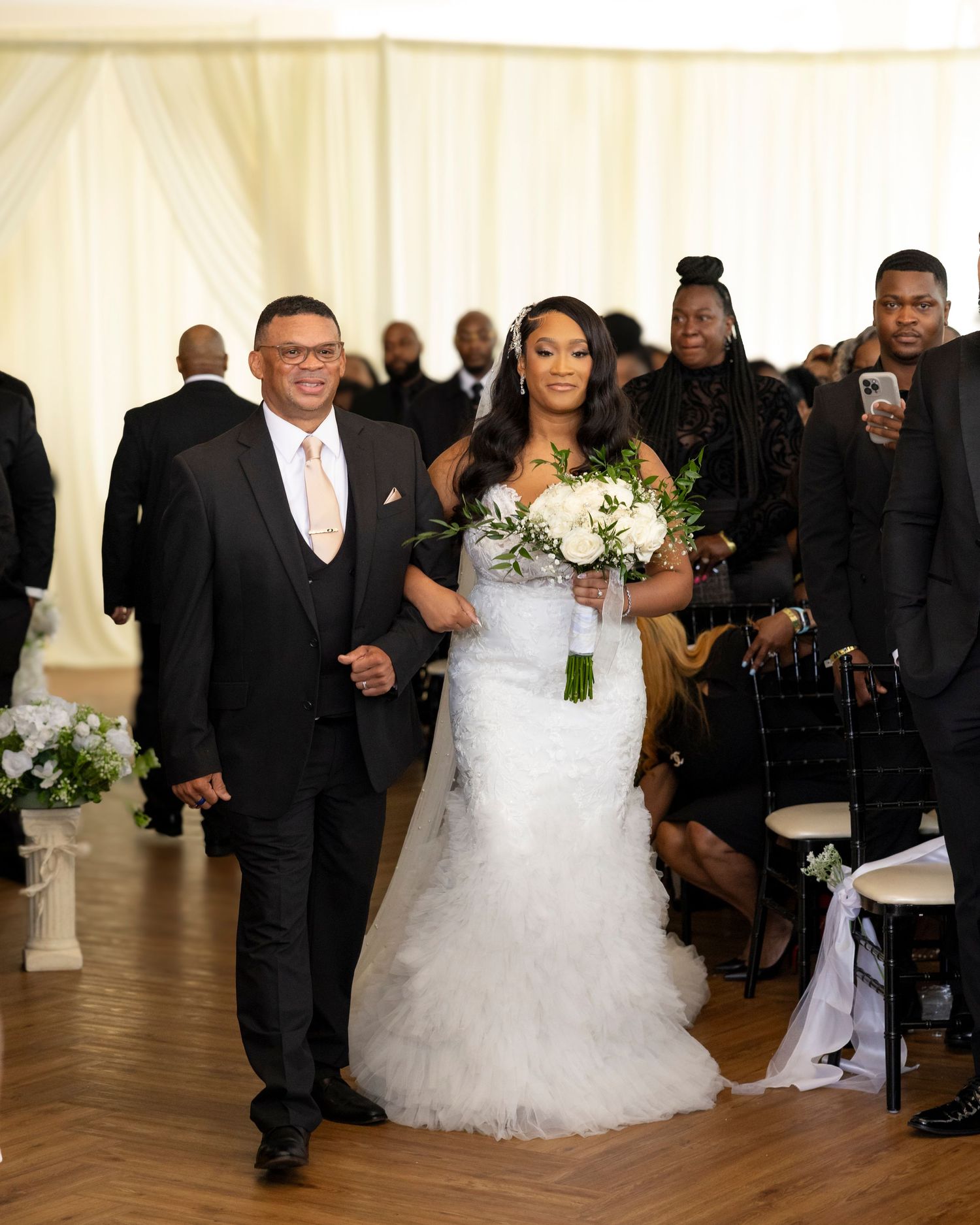 A bride and groom walk down the aisle at an elegant indoor wedding ceremony with white draped decor.