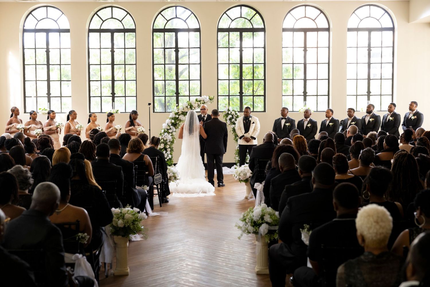 Indoor wedding ceremony with elegant arched windows, guests seated in rows, and floral arrangements in a bright venue.