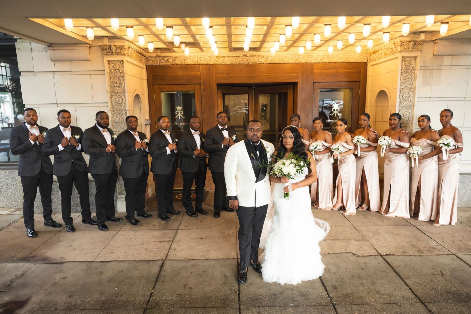 Wedding party in formal black suits and blush pink bridesmaid dresses pose elegantly outside a venue with illuminated ceiling.
