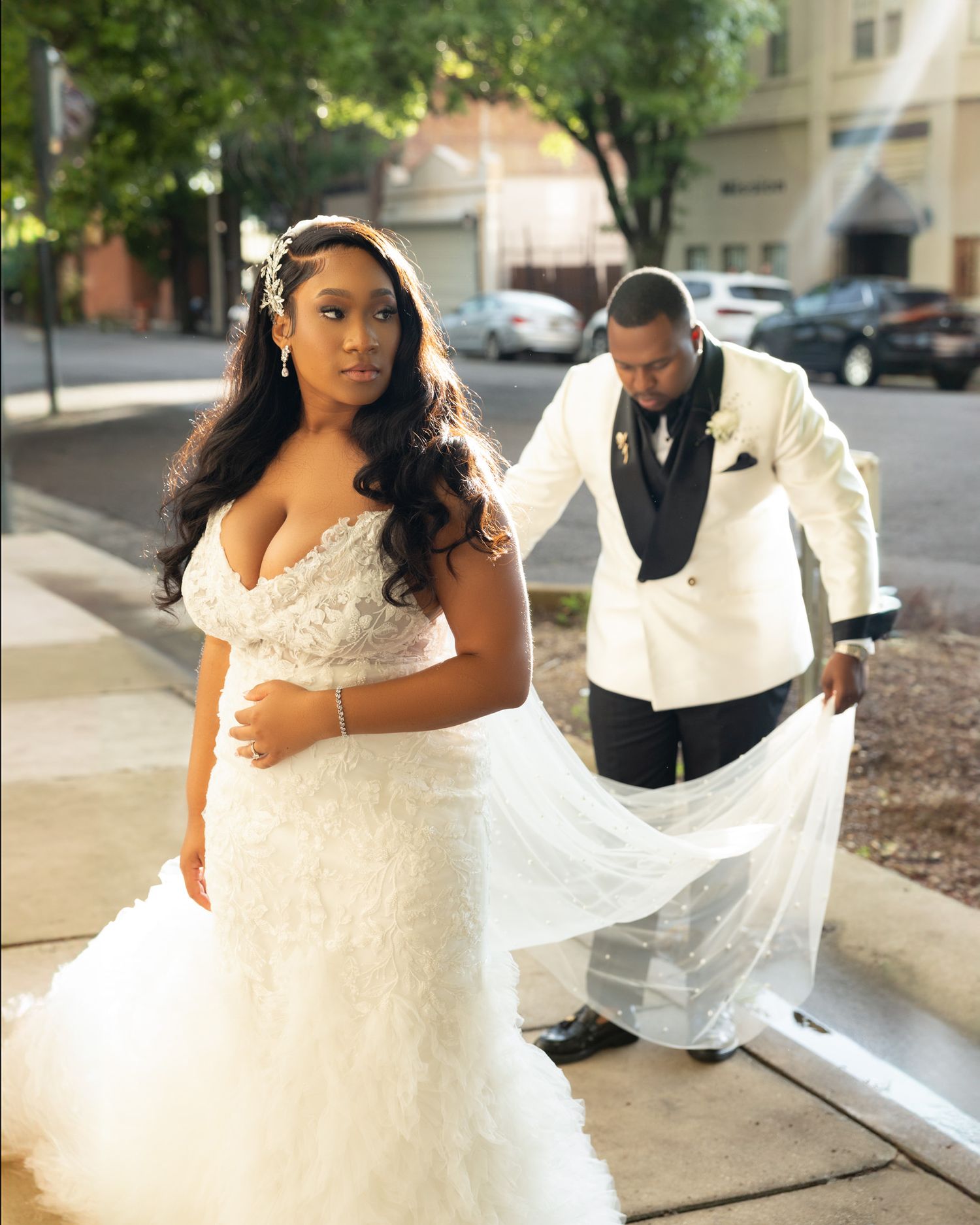 A couple poses outdoors on their wedding day, with the bride in a white lace gown and flowing veil.