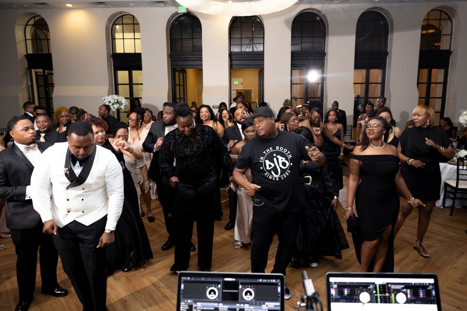 Party guests dance and celebrate on a dance floor in front of DJ equipment at an elegant venue with arched windows.