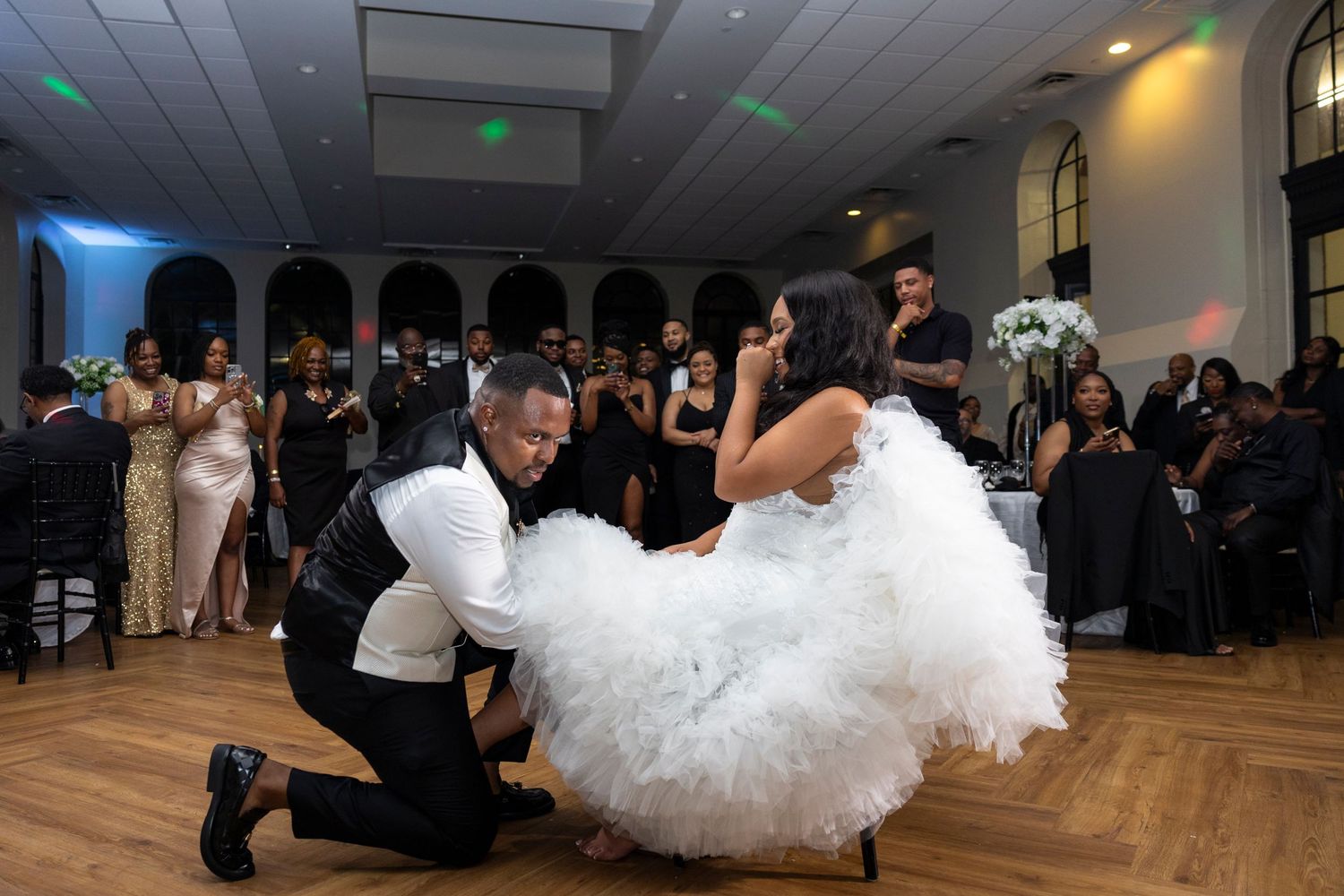 A couple performs a playful dance move during a wedding reception on a wooden dance floor surrounded by guests.