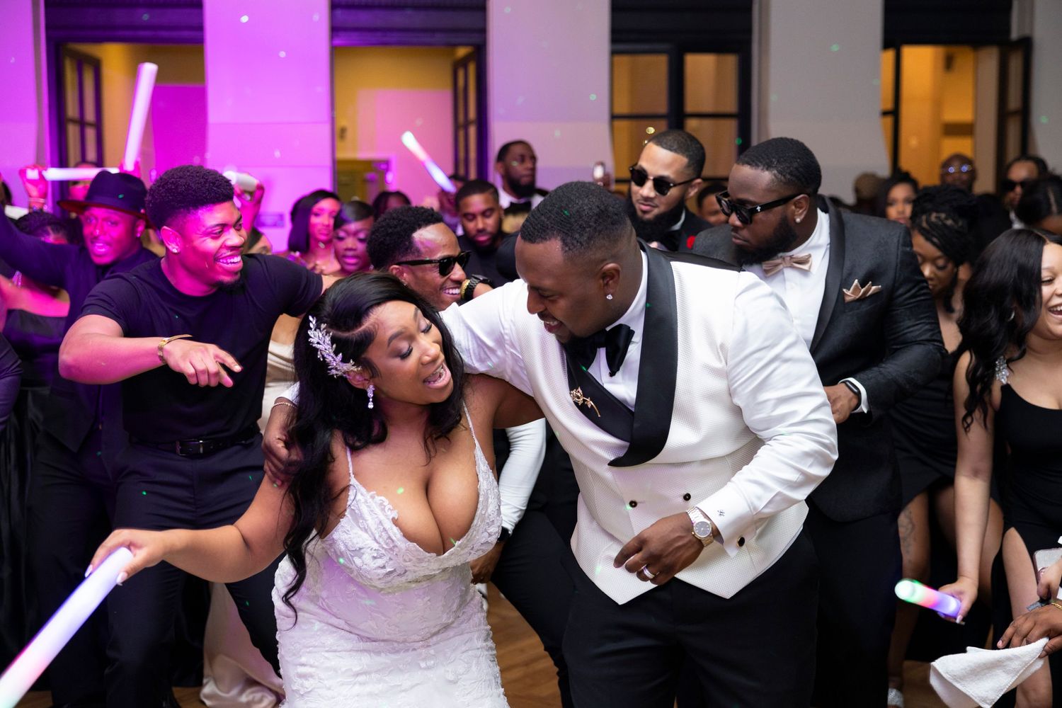 Wedding party dances energetically on the dance floor under purple lighting at a reception celebration.