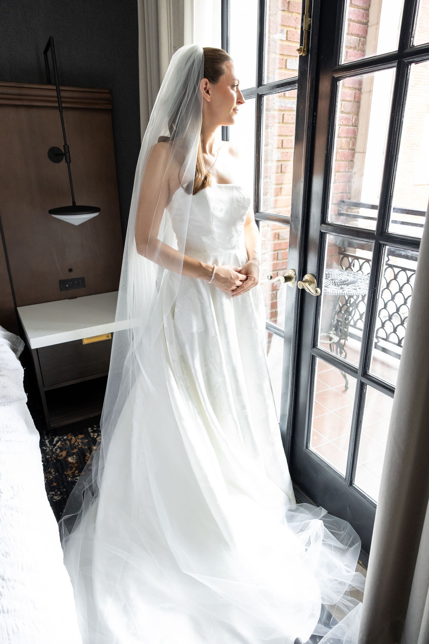 Bride in white gown and veil stands by window in hotel room on wedding day.