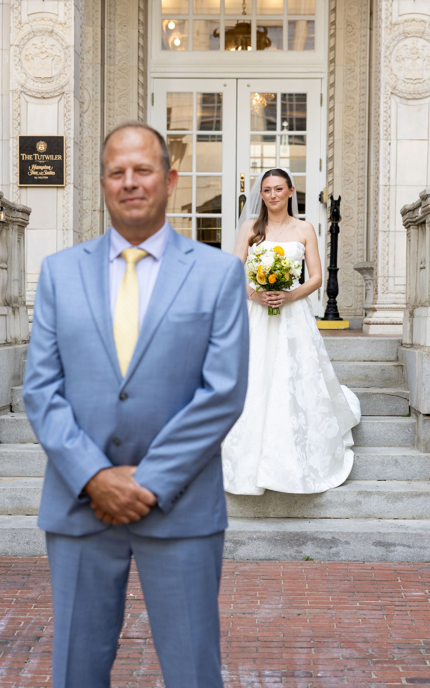 Bride in white gown holding yellow bouquet stands on steps behind man in light blue suit during wedding first look.
