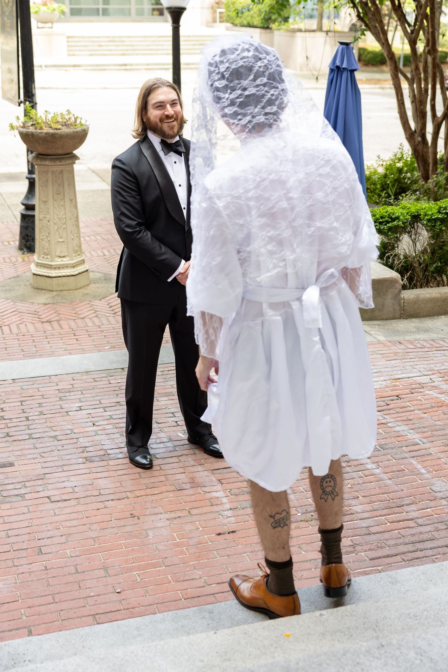 Groom in tuxedo smiles at tattooed person in white wedding veil and robe during outdoor wedding ceremony.