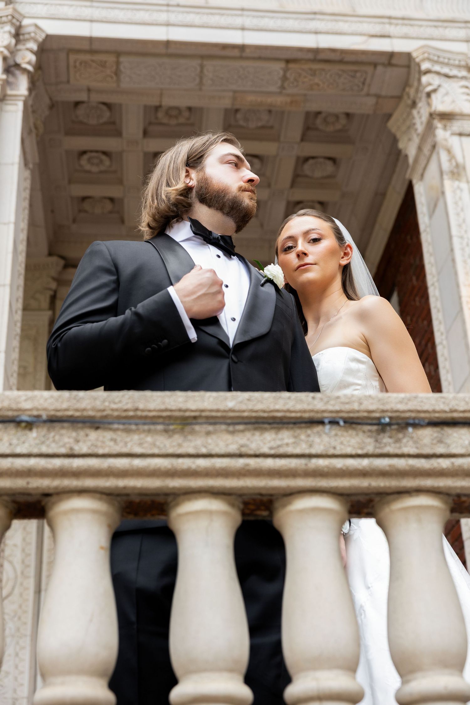 Elegant bride and groom pose on a stone balcony, groom in black tuxedo, bride in white gown with veil.