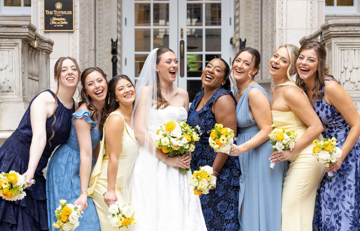 Bride and bridesmaids in blue and yellow dresses laughing together outside elegant venue.