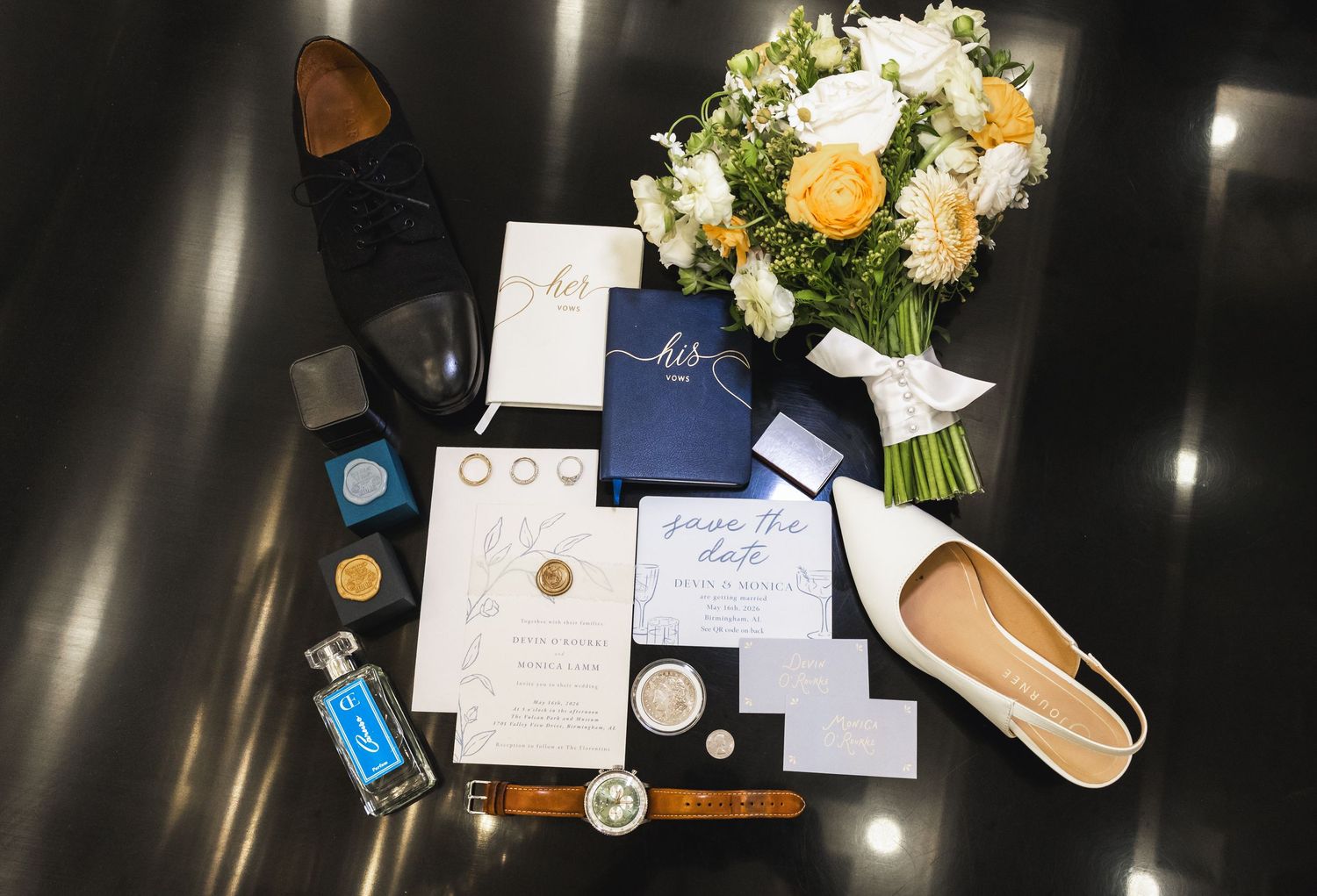 Elegant wedding flat lay featuring navy invitations, floral bouquet, shoes, rings, and accessories on dark surface.