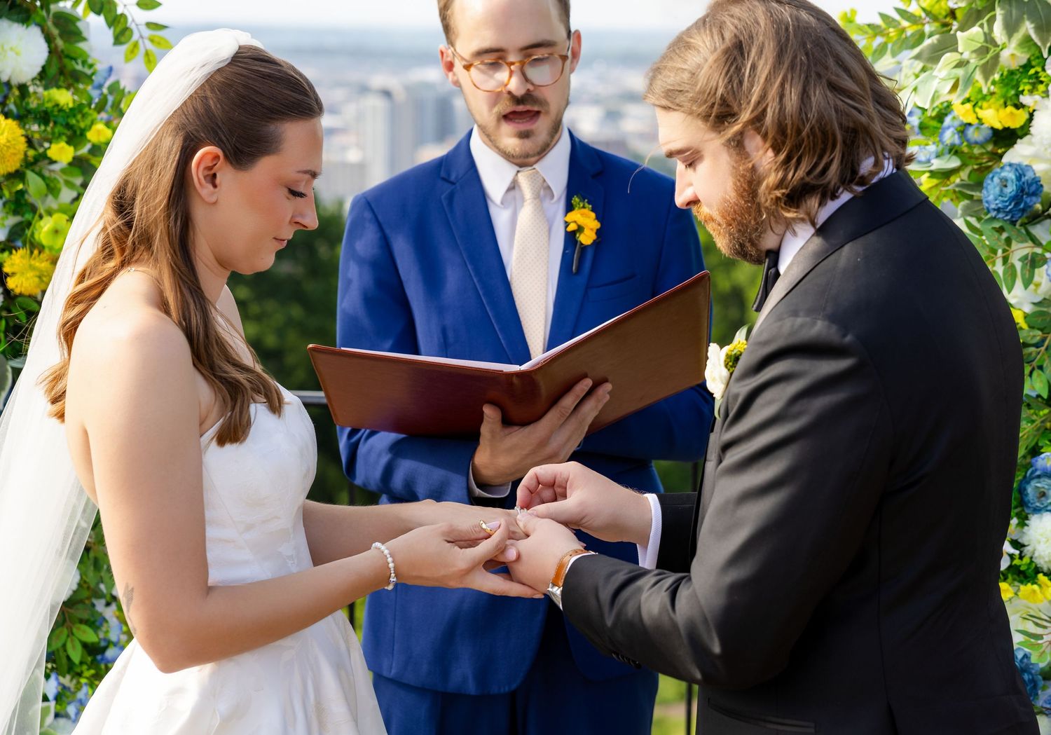 Bride and groom exchange rings during outdoor wedding ceremony with officiant.