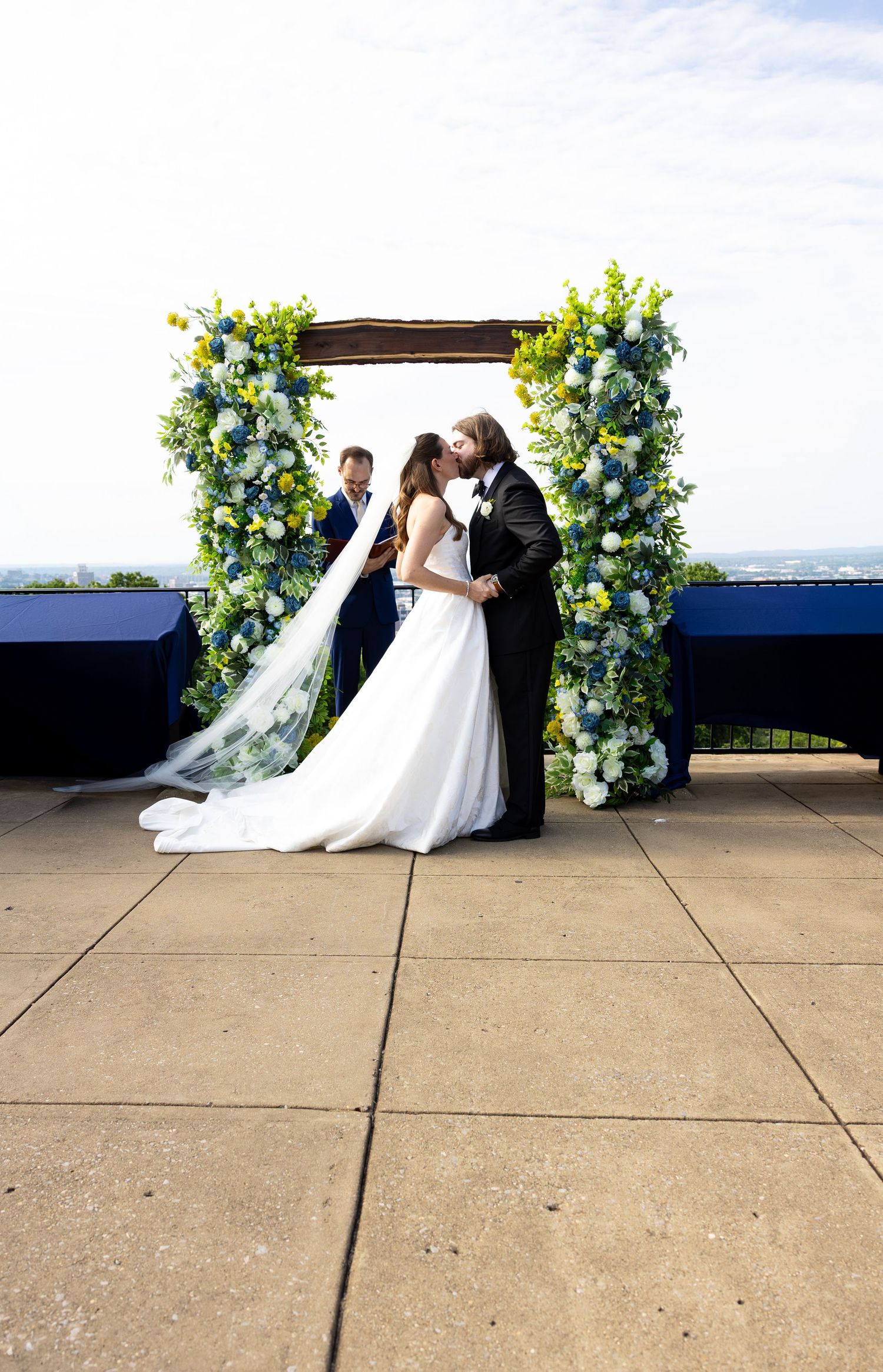 Bride and groom share first kiss under floral arch with blue and yellow flowers at outdoor ceremony.