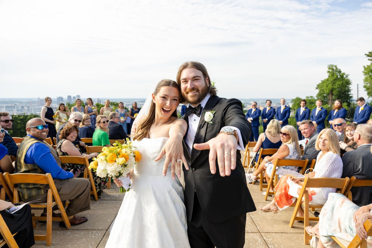 Newlyweds show off wedding rings at outdoor ceremony with guests seated behind them.
