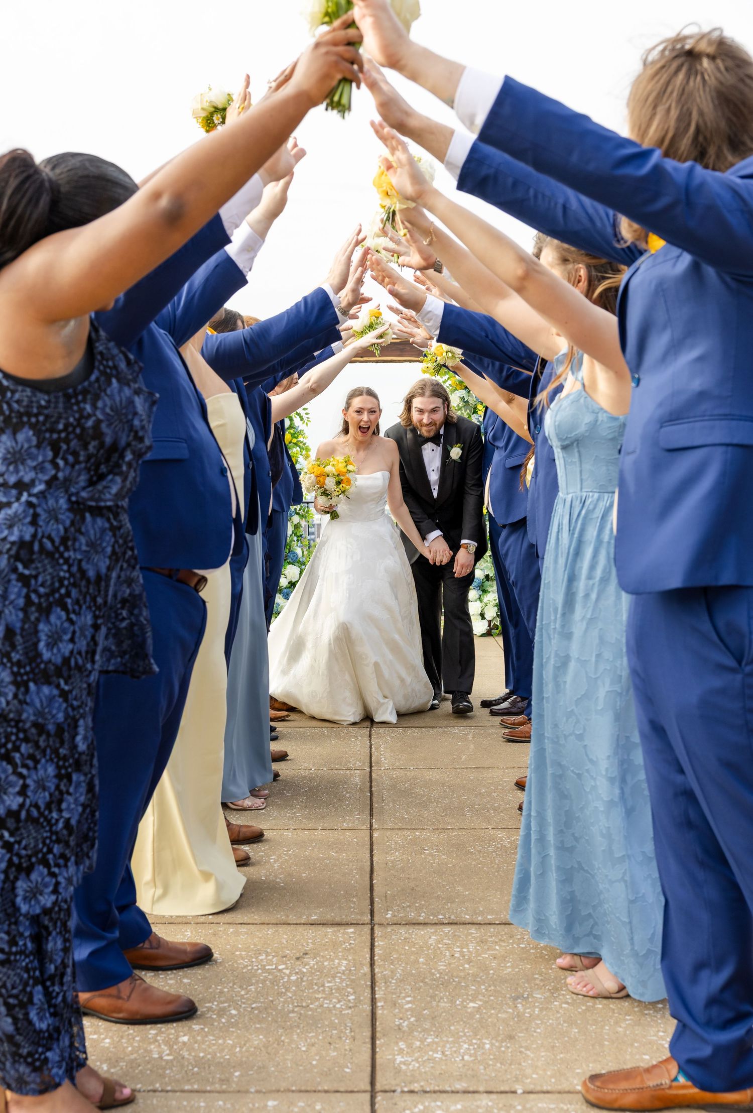 Wedding party forms an arch with bouquets as bride and groom walk through in blue and white attire.