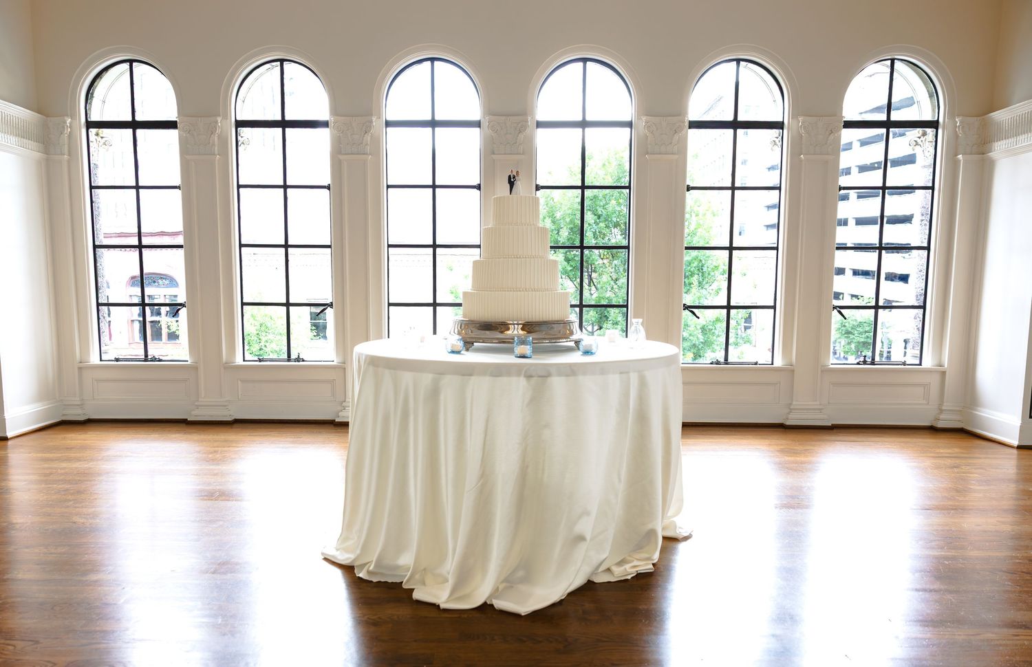 Elegant white wedding cake displayed on draped table in bright ballroom with arched windows.