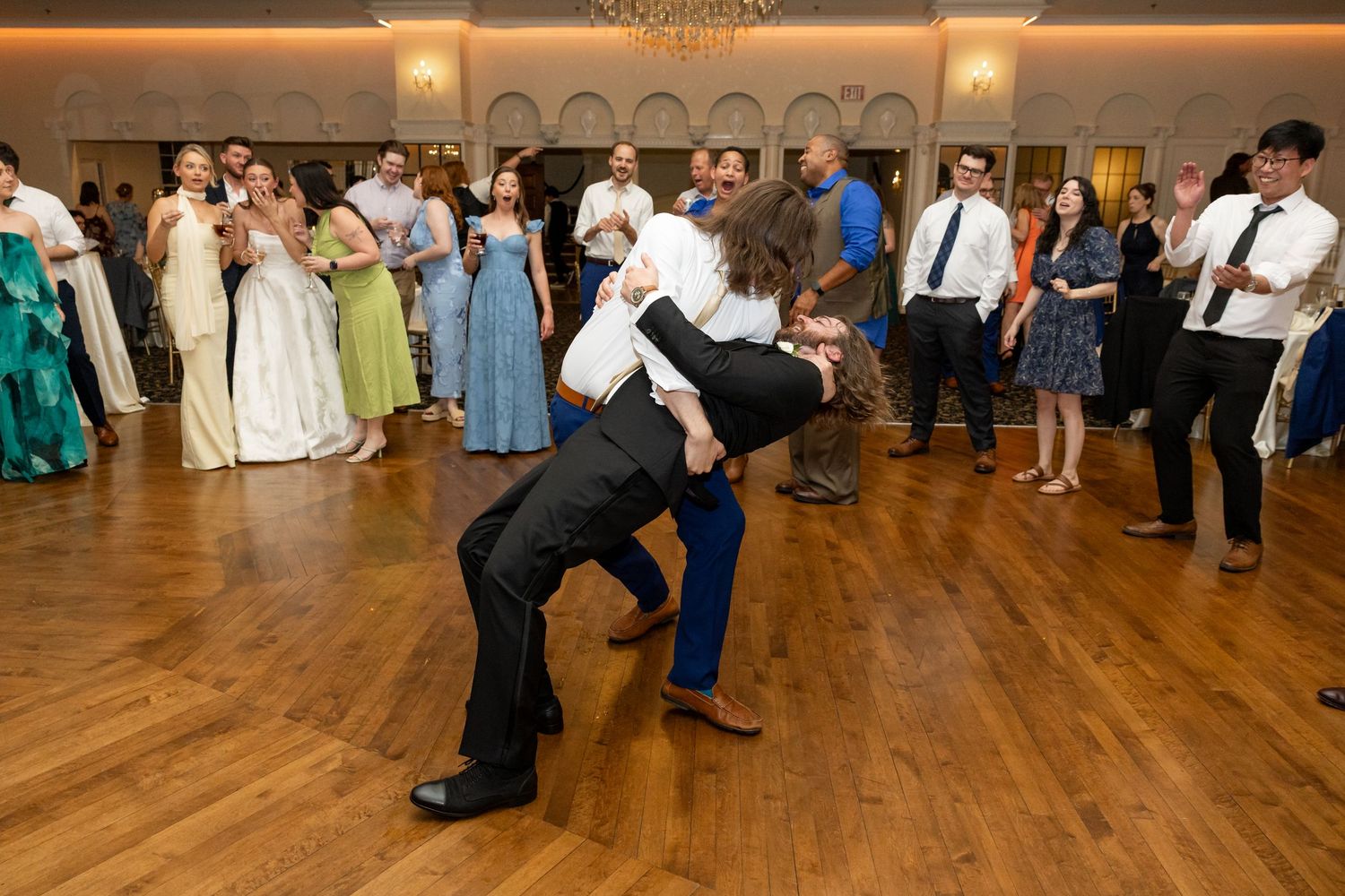 Wedding guests watch as couple shares energetic dance move on ballroom floor during lively wedding reception.