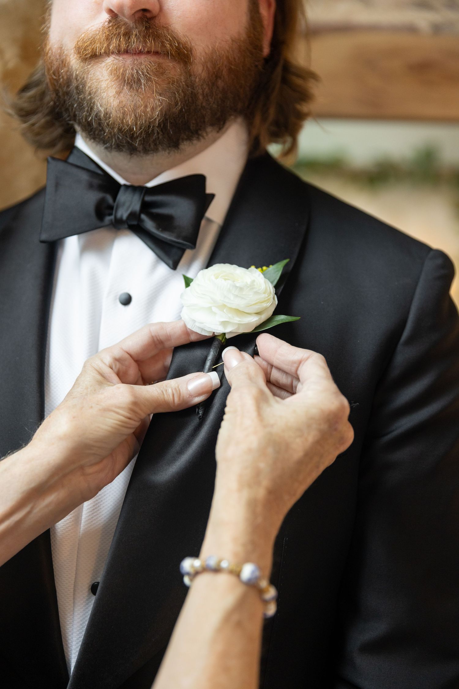 Hands pinning a white rose boutonniere on a bearded groom in a black tuxedo with bow tie.