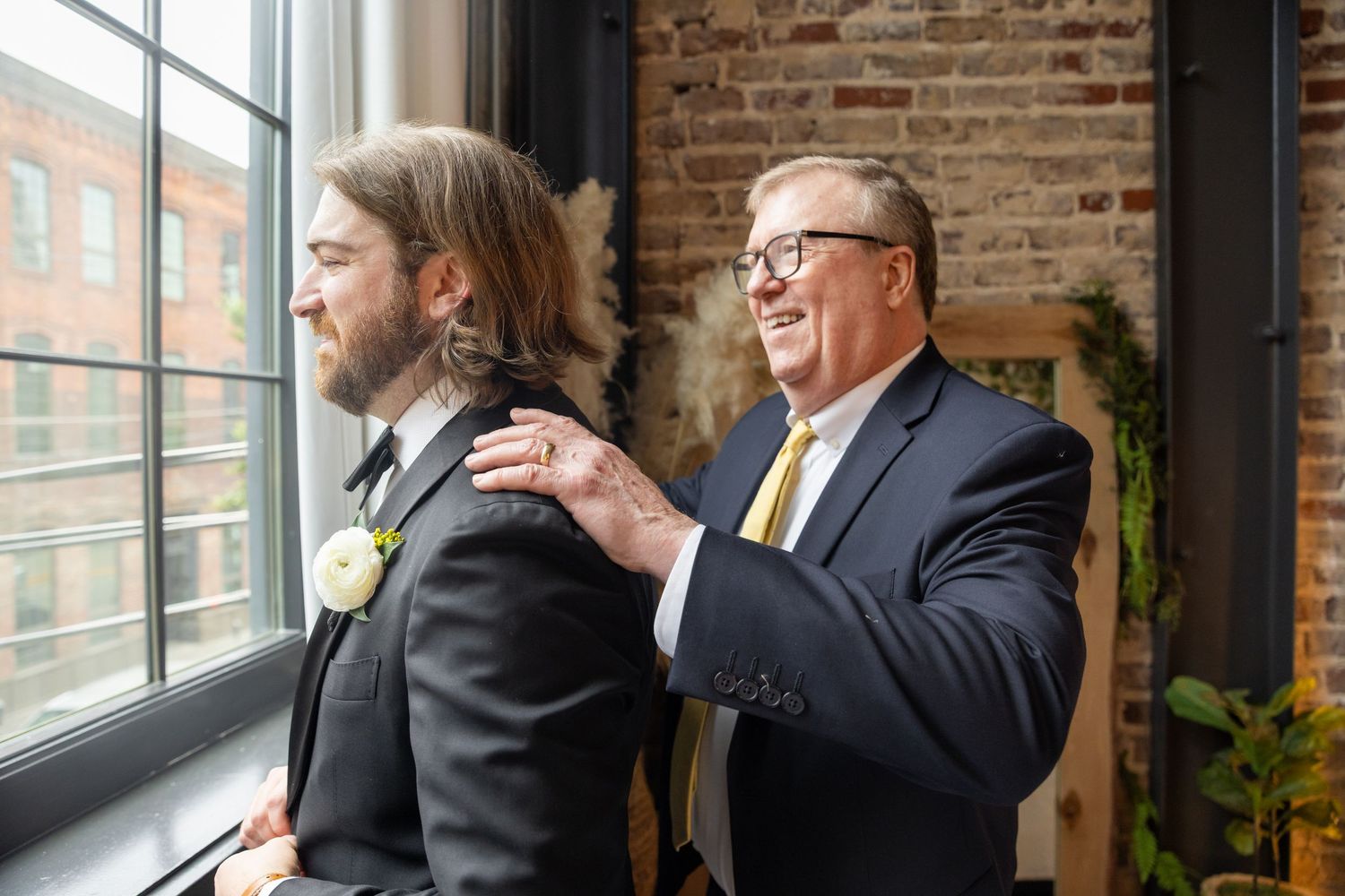 Father adjusting groom's suit jacket near window before wedding ceremony in rustic brick venue.