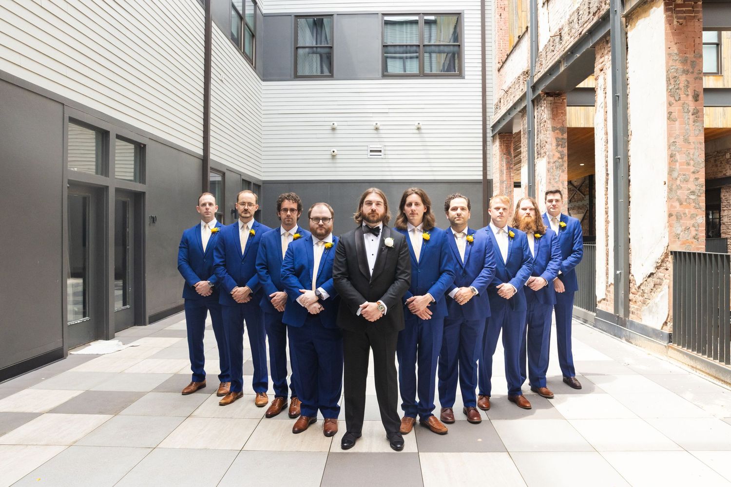 Group of men in matching blue suits posing in a courtyard, led by a bearded man in a black suit.