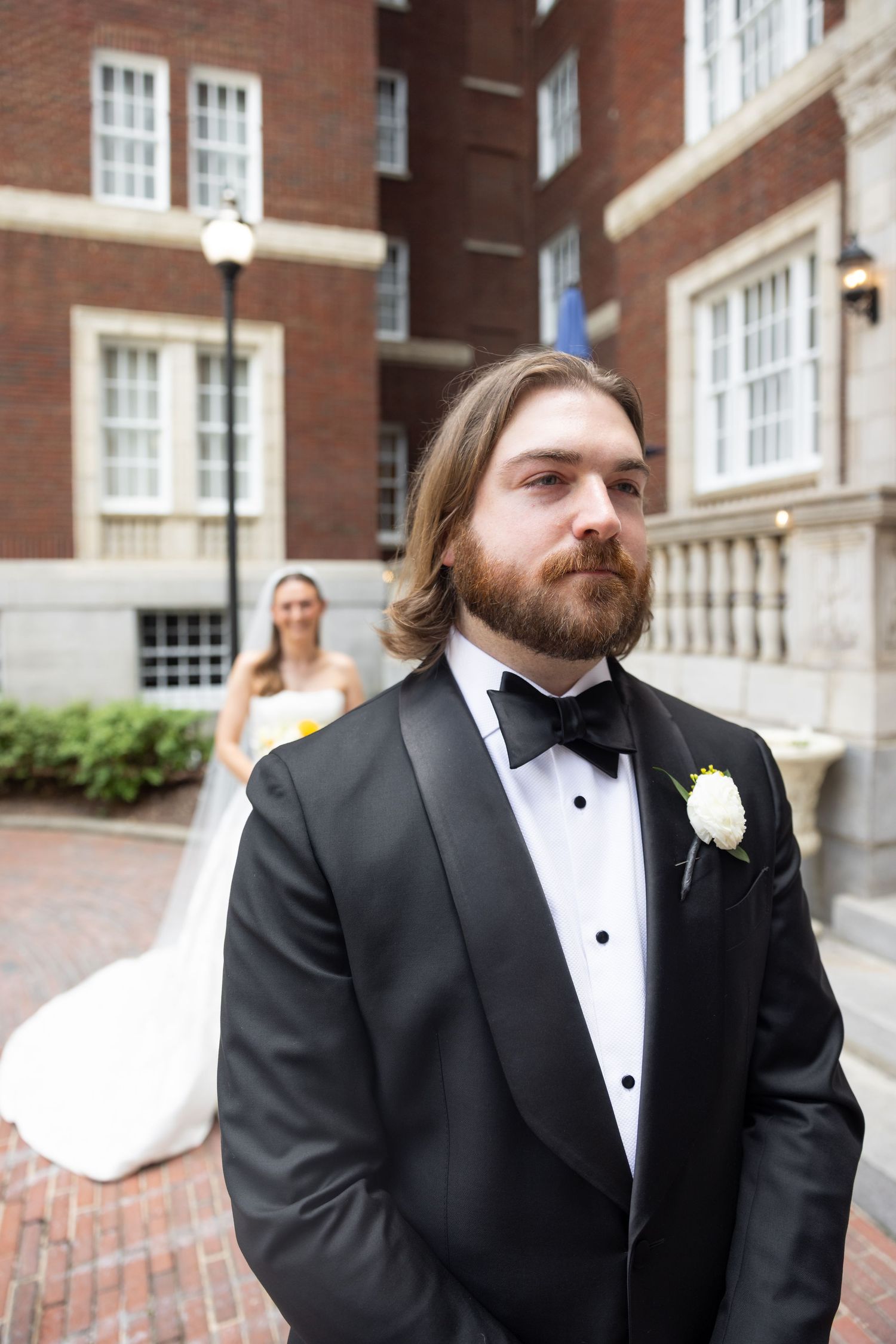 Groom in black tuxedo with white boutonniere stands outdoors, bride in white gown visible in background near brick building.