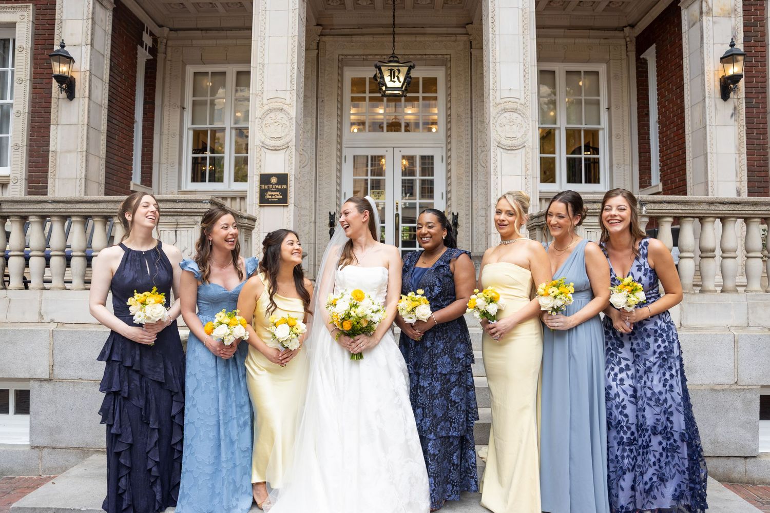 Bride in white gown with six bridesmaids in blue and yellow dresses holding sunflower bouquets outside elegant brick building.