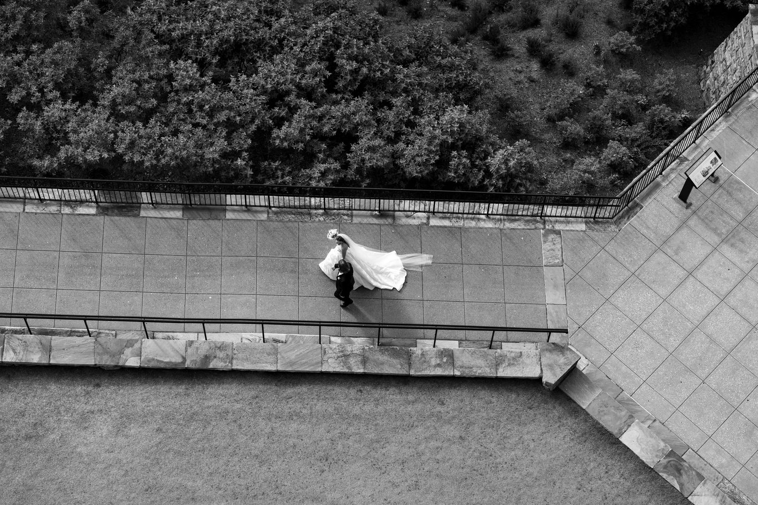 Aerial black and white photo of a bride in a flowing gown walking along a paved terrace path near lush greenery.