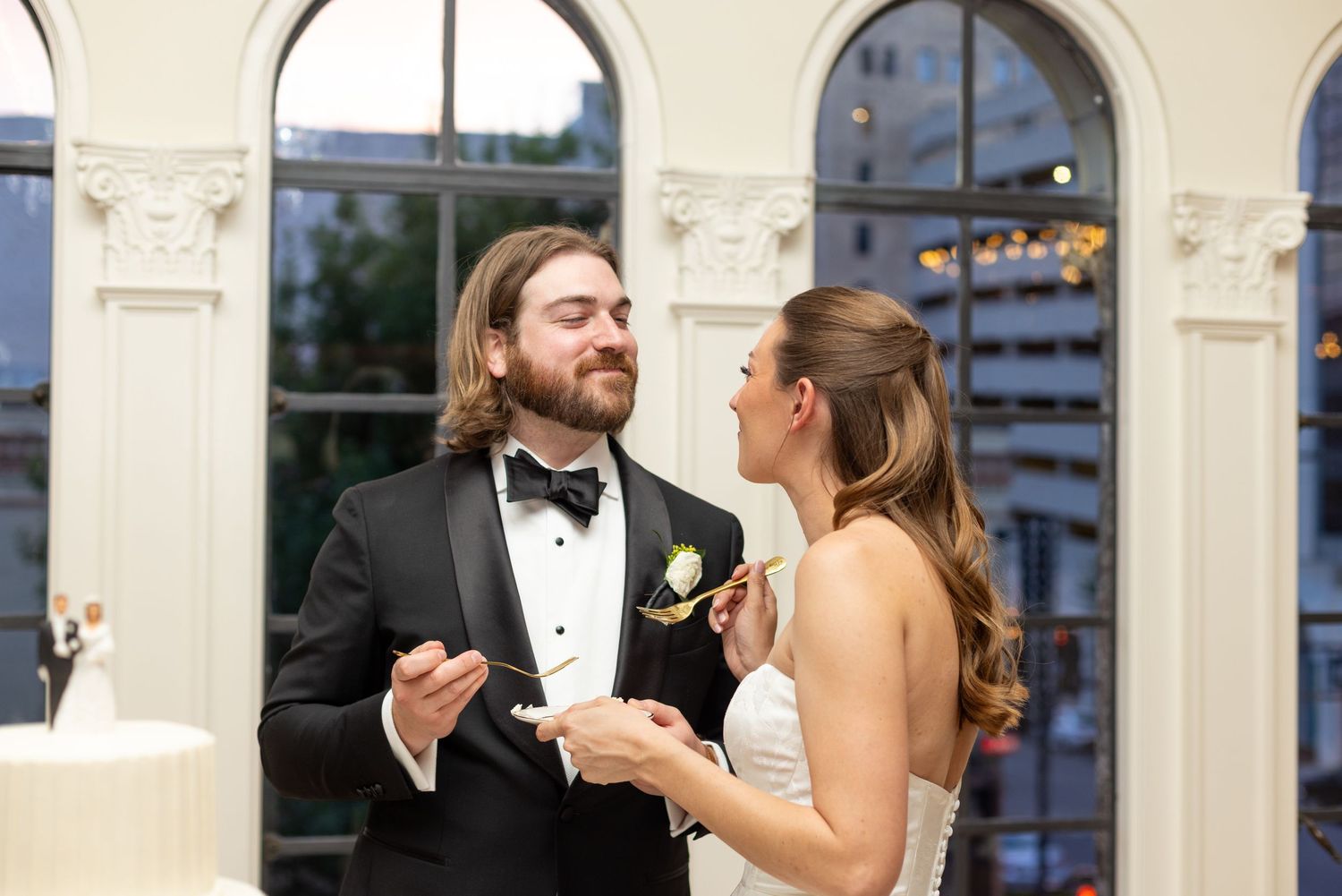 Bride and groom share wedding cake during elegant indoor reception, groom in tuxedo, bride in white gown.