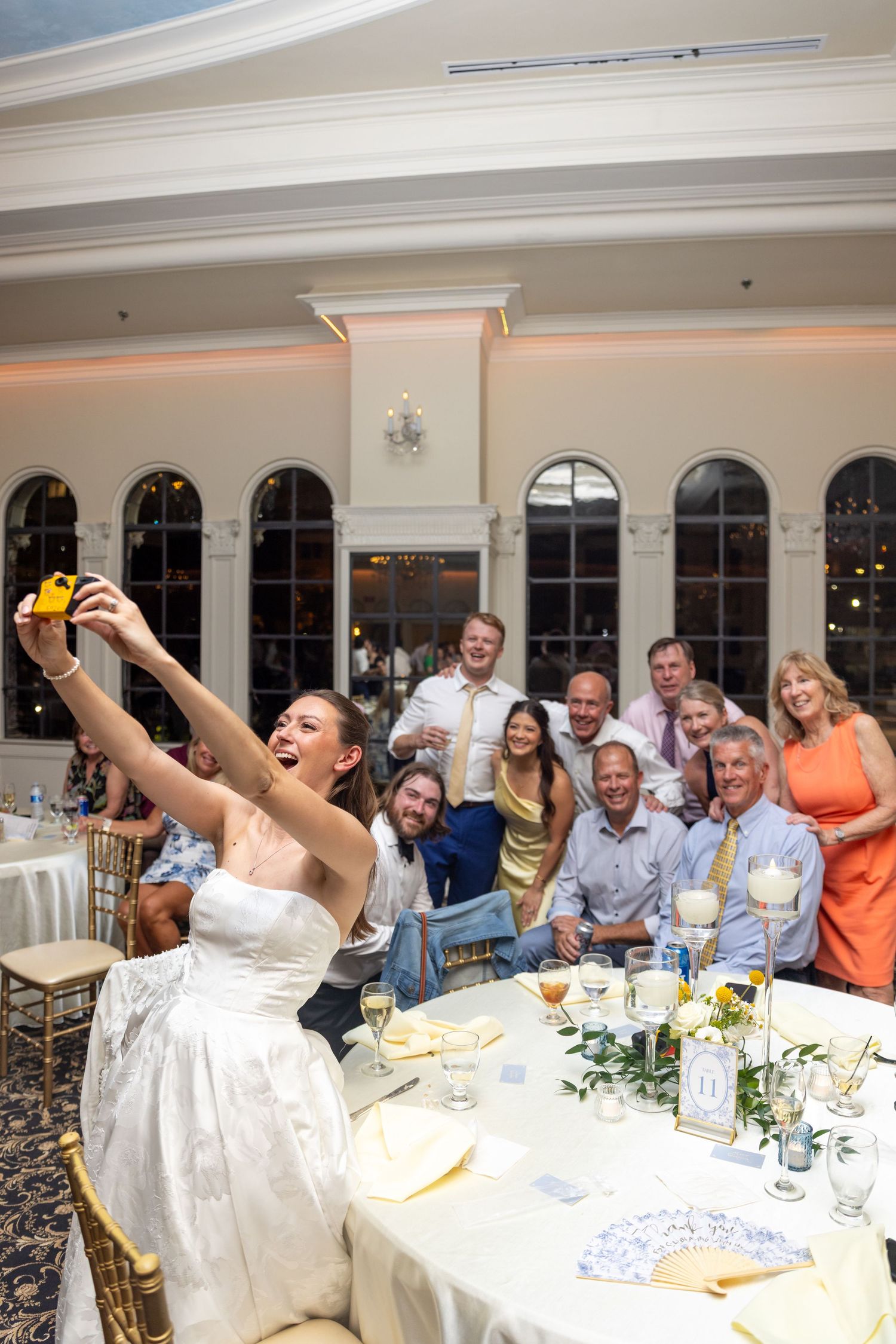 Bride takes a selfie with wedding guests at a elegant reception venue, capturing joyful smiles around a decorated table.