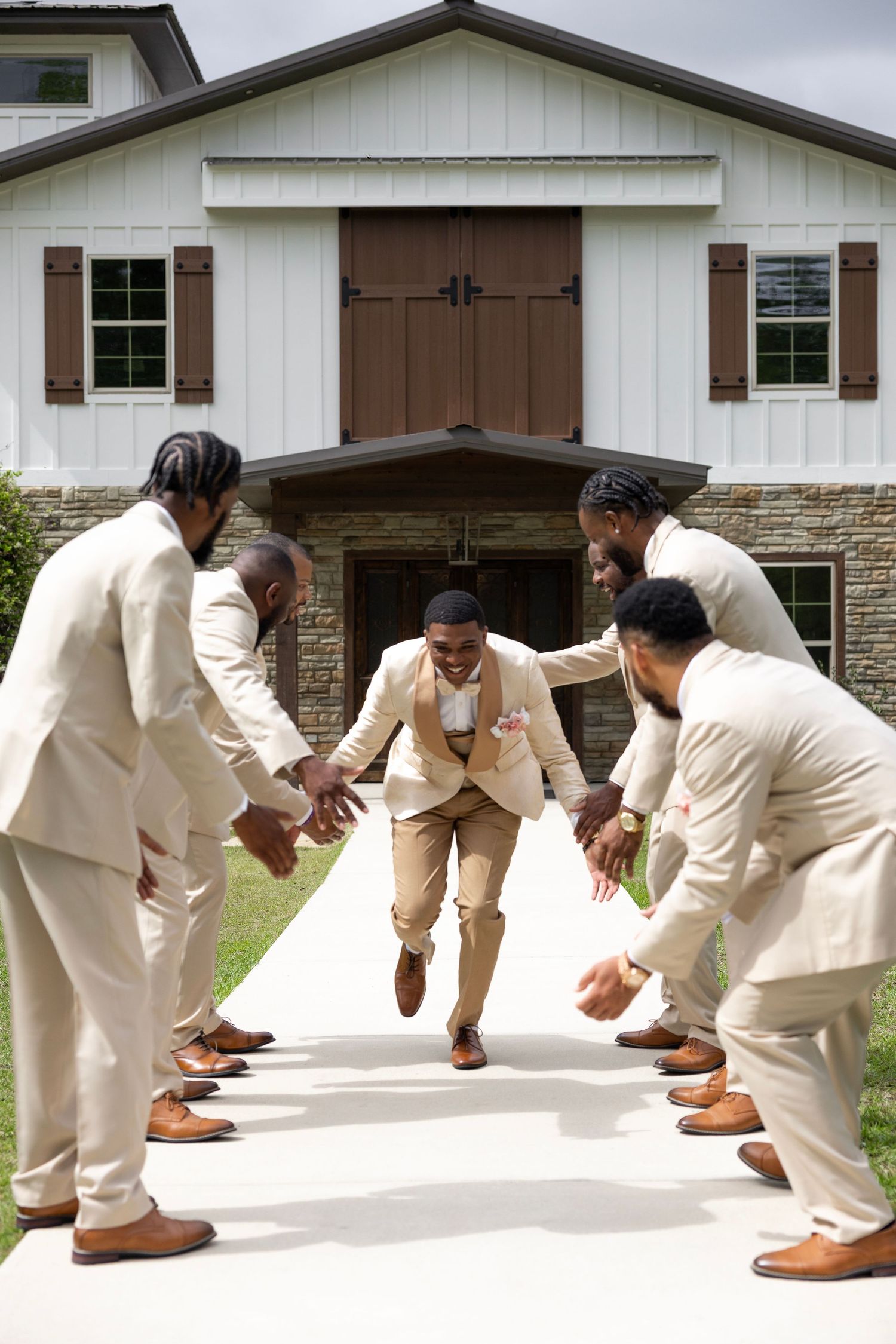 Groomsmen in tan suits celebrate groom running through a tunnel of hands outside a white barn venue.