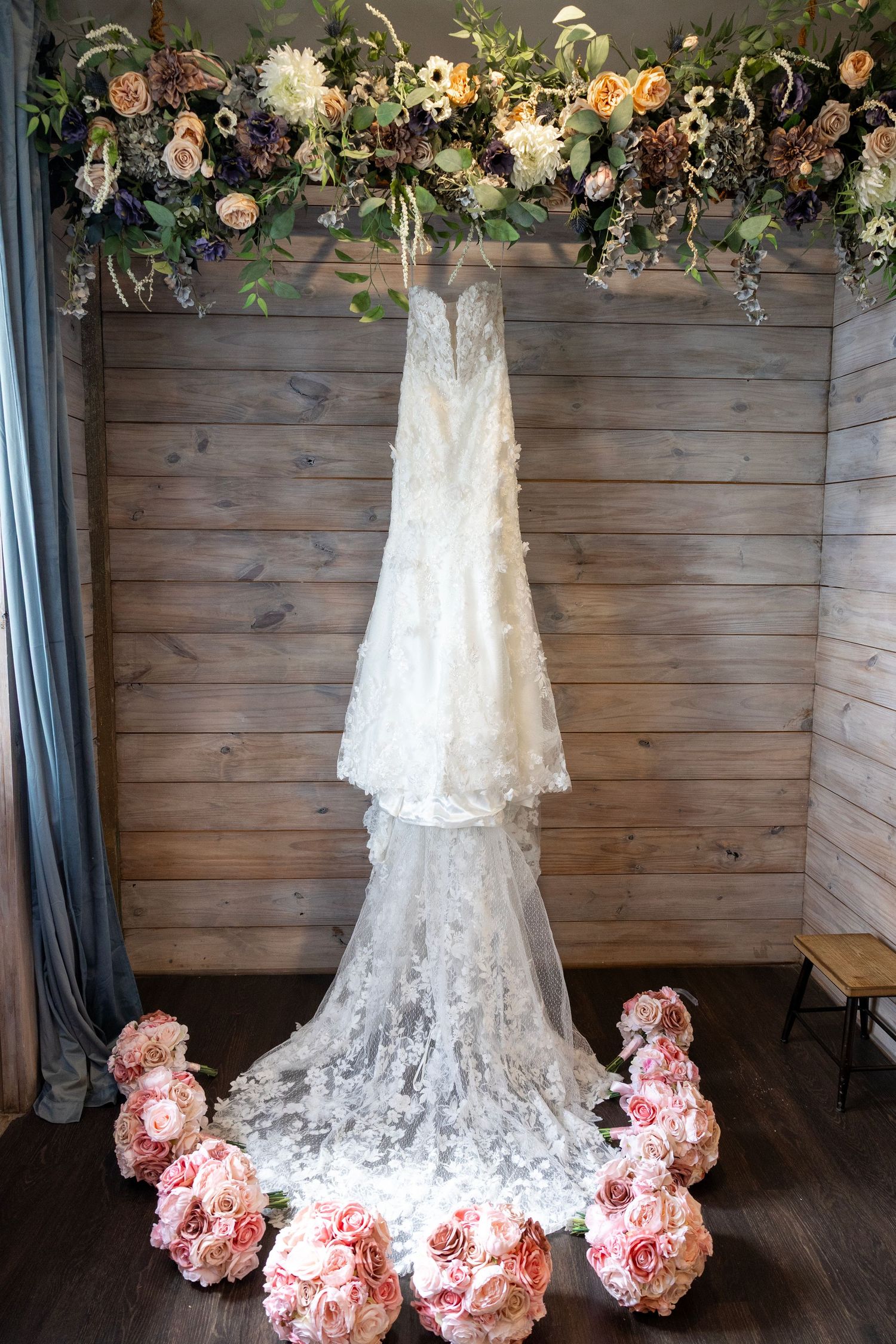 White lace wedding dress hanging above pink rose bouquets against rustic wood wall with floral arrangement overhead.