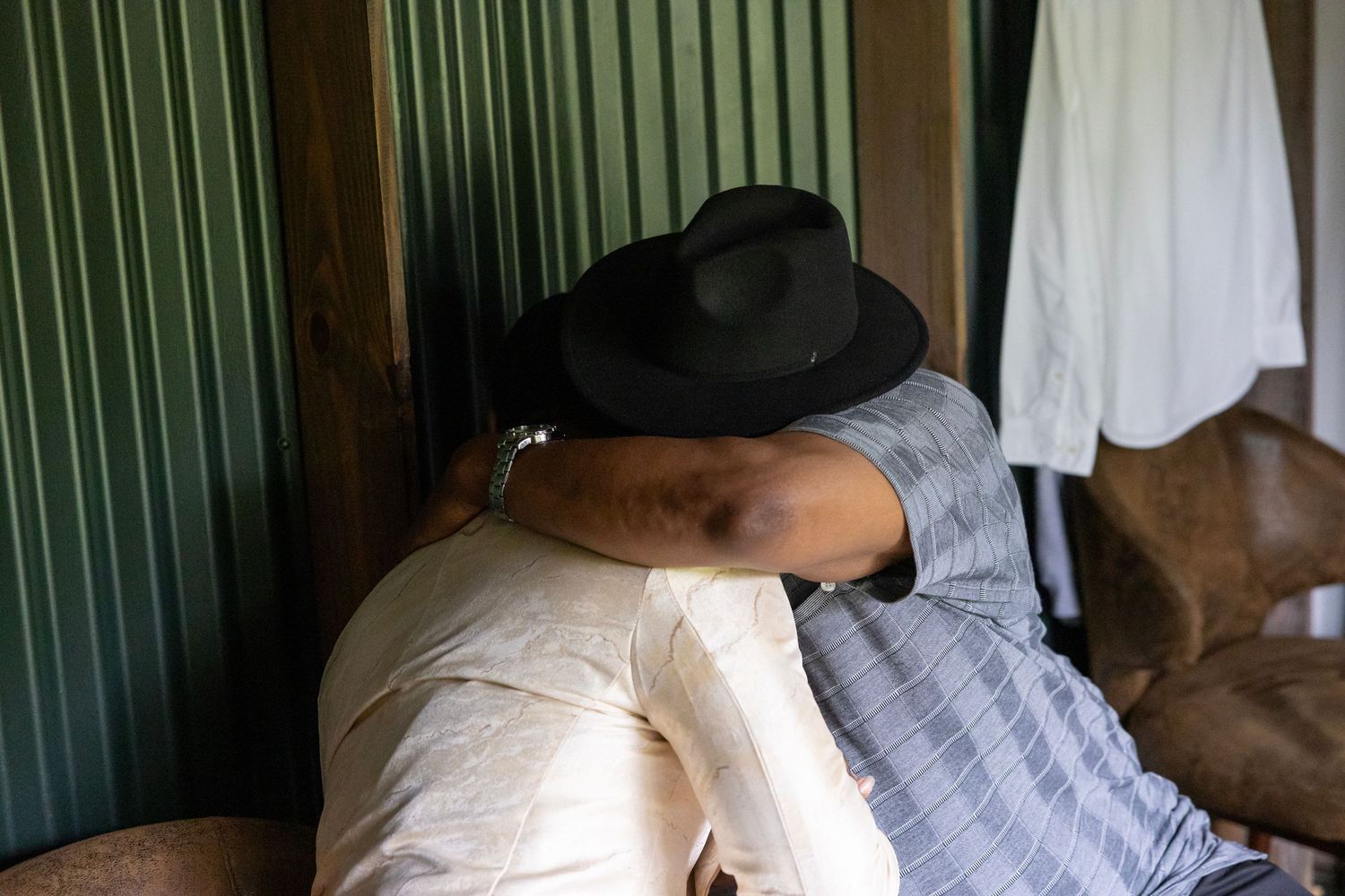 Two people embracing warmly indoors, one wearing a black cowboy hat, near green walls and hanging white clothing.