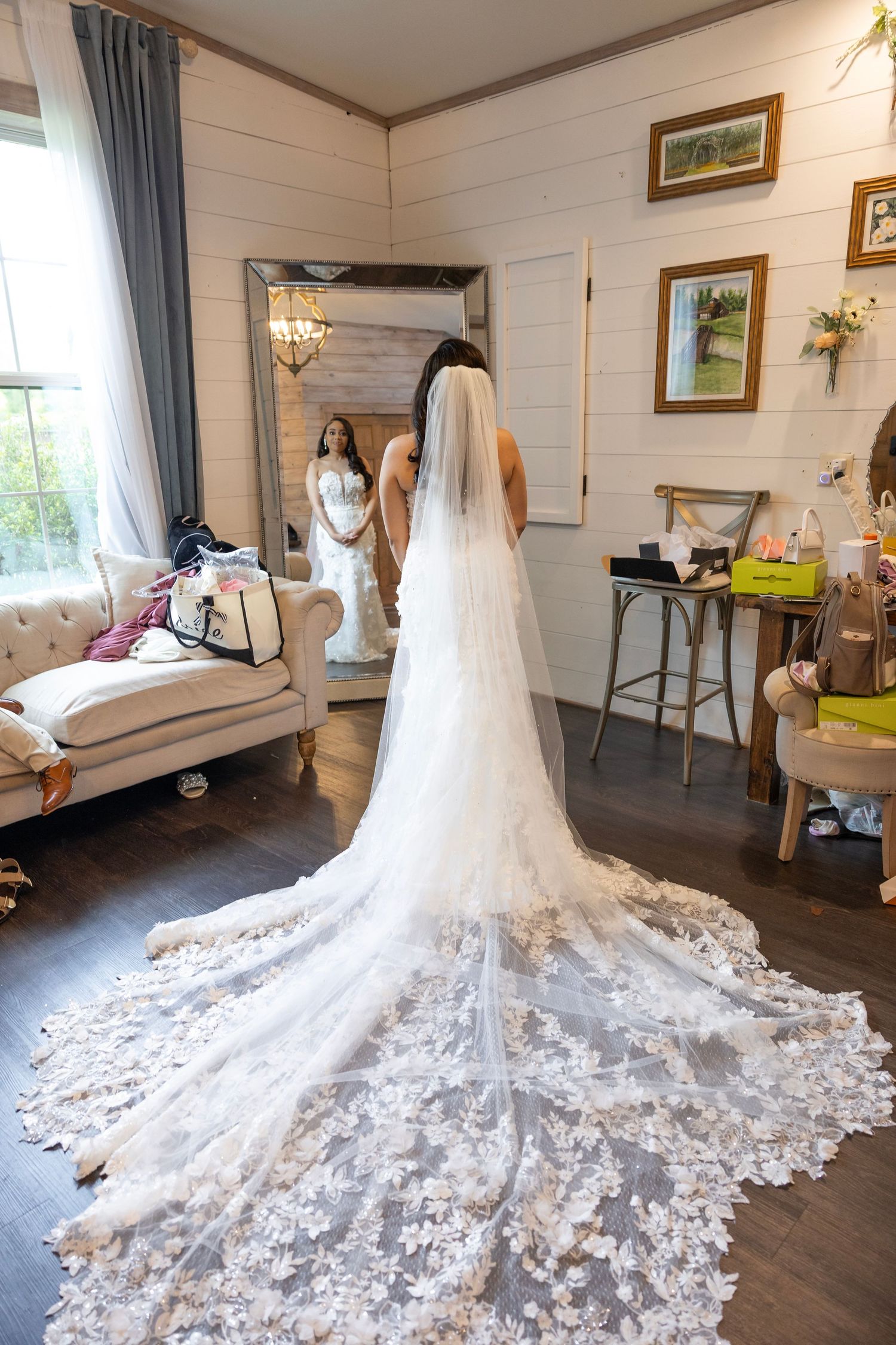 Bride in elegant lace wedding gown with long floral veil viewed from behind, reflected in mirror in bridal suite.