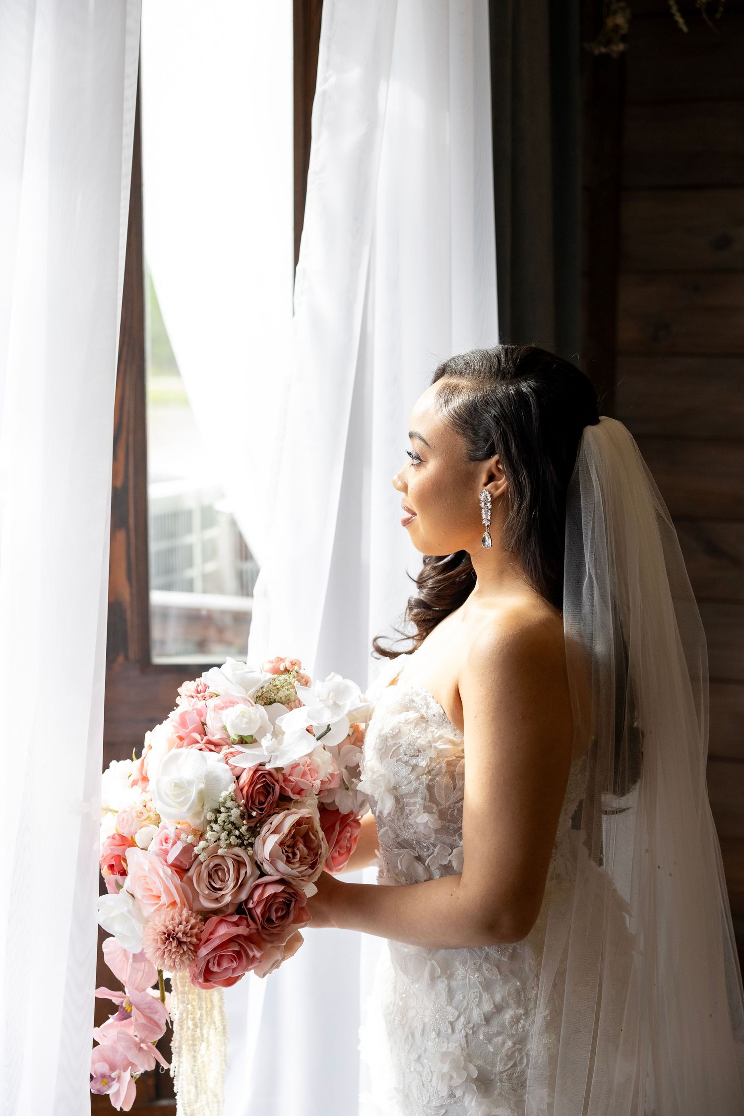 Bride in lace gown holding pink bouquet gazes out window with white curtains on her wedding day.