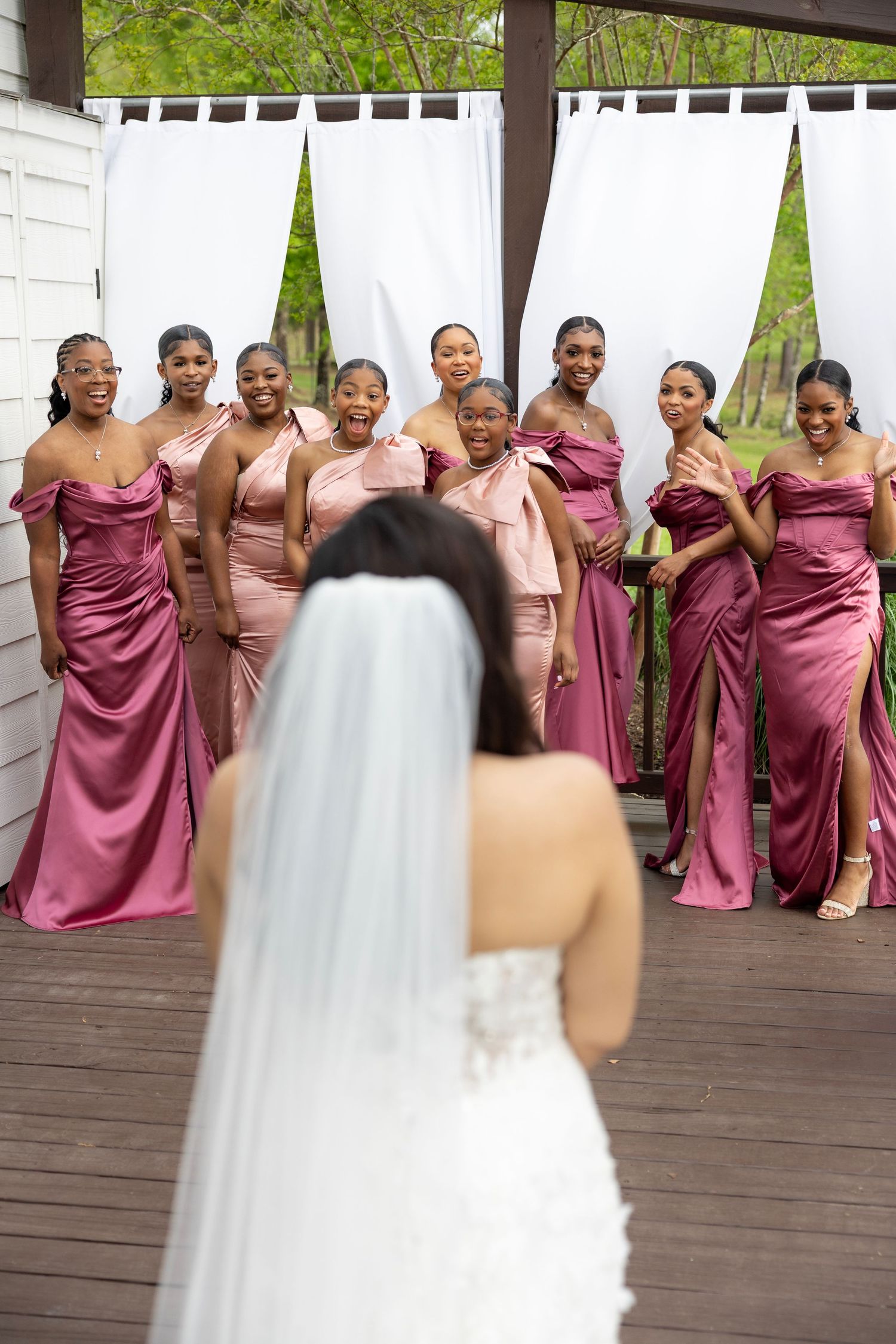 Bride in white gown and veil reveals herself to bridesmaids in pink satin dresses on a wooden deck.