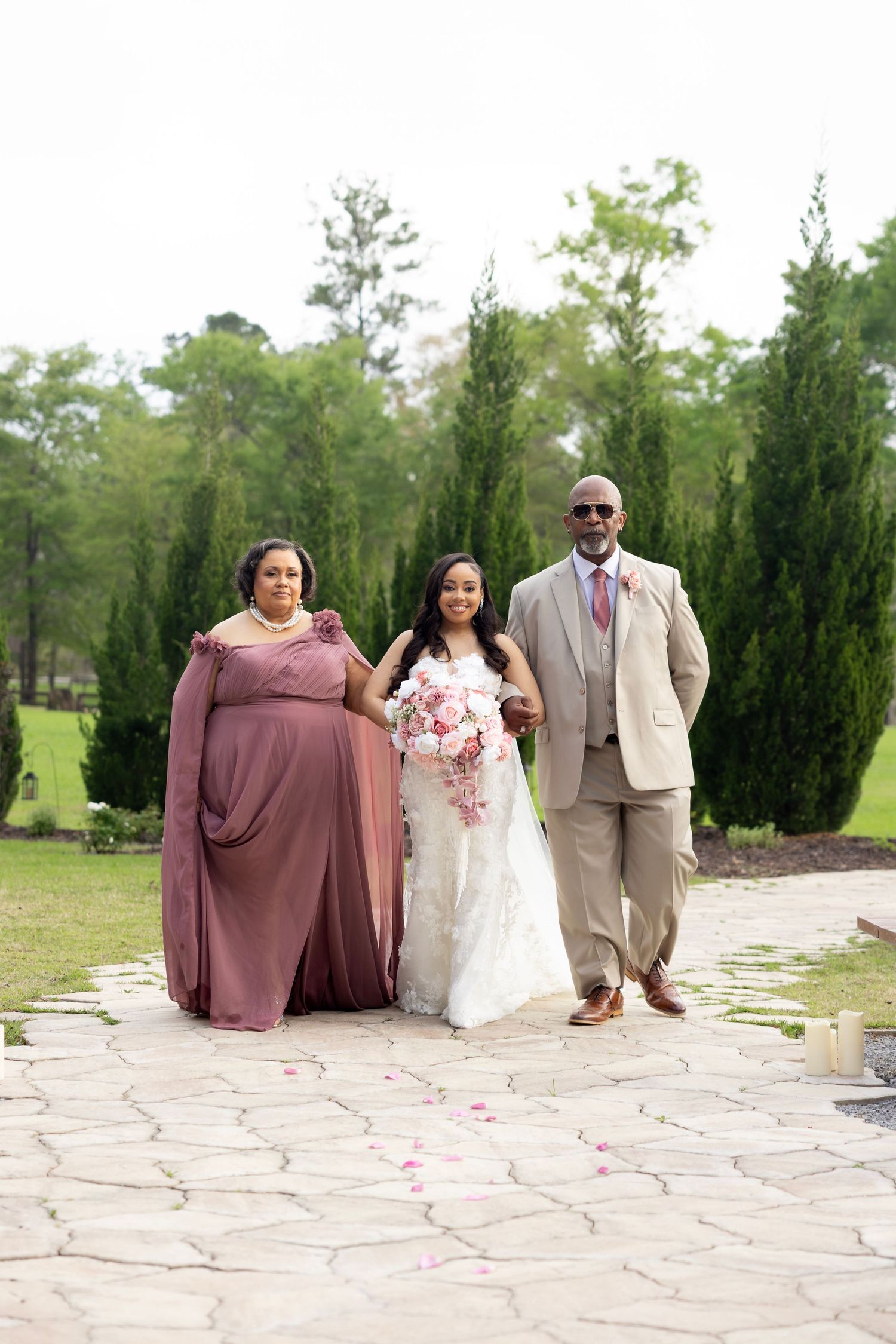 Bride in white gown walks with parents on stone path, woman in mauve dress and man in tan suit, outdoor wedding ceremony.
