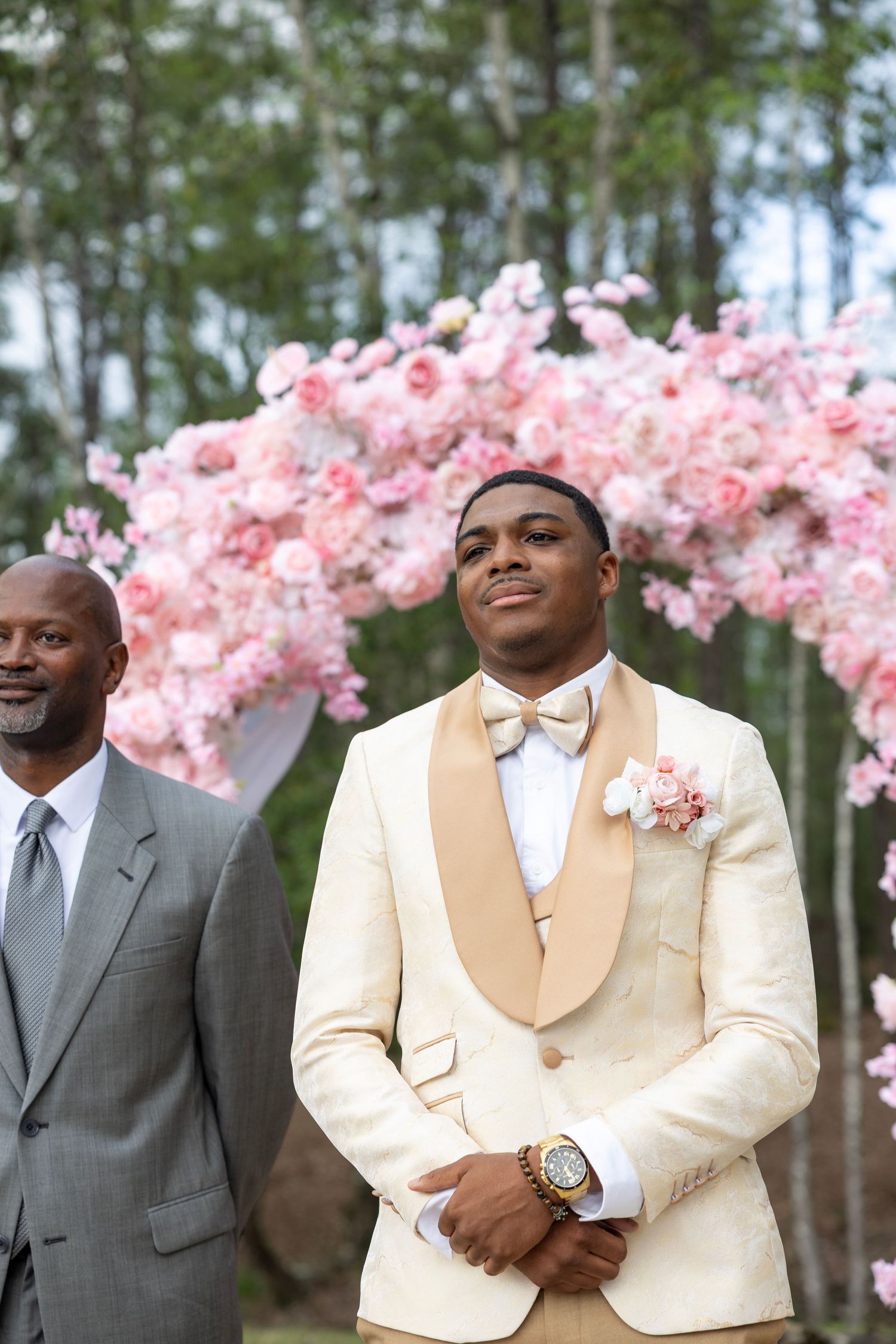 Groom in cream tuxedo with pink boutonniere stands at outdoor wedding altar adorned with pink floral arch.