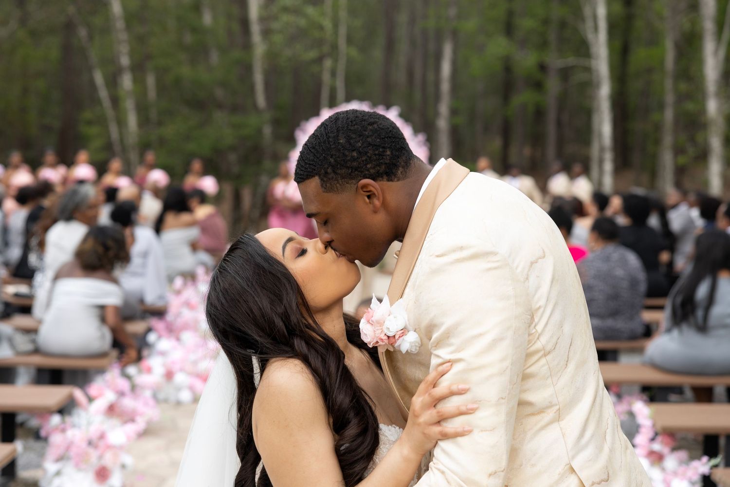 Couple shares first kiss at outdoor woodland wedding ceremony with pink floral decorations and guests seated on benches.