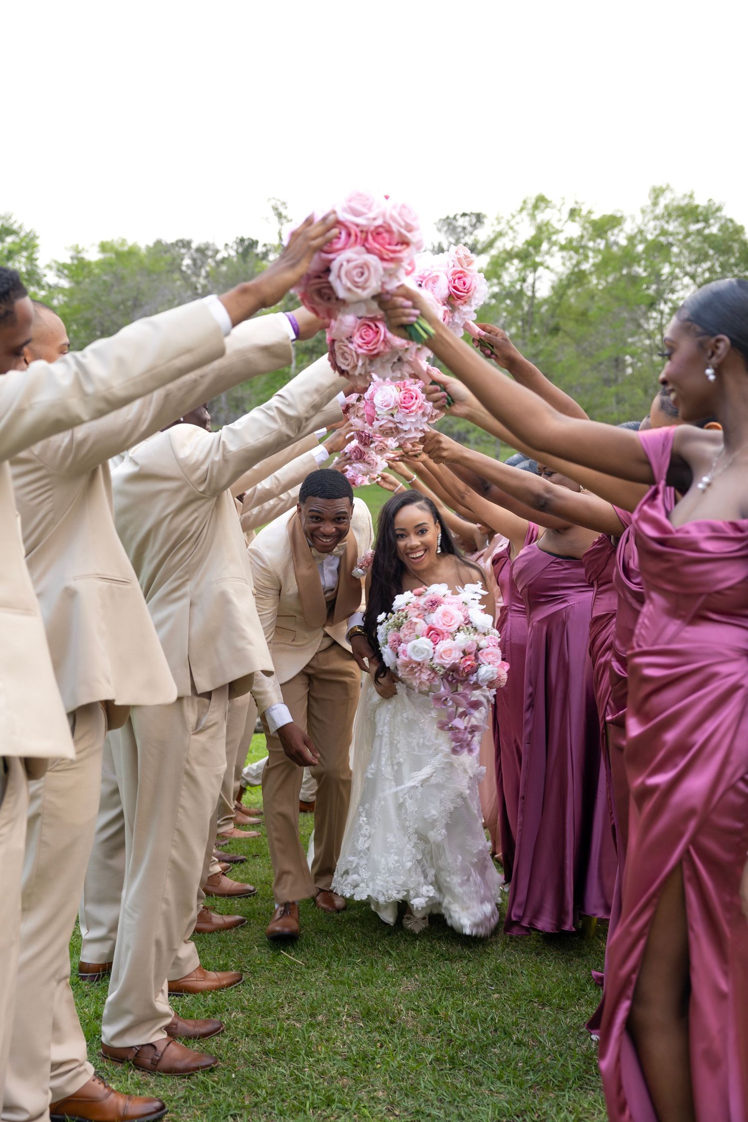 Bride and groom walk through floral arch tunnel made by wedding party in pink dresses and tan suits outdoors.