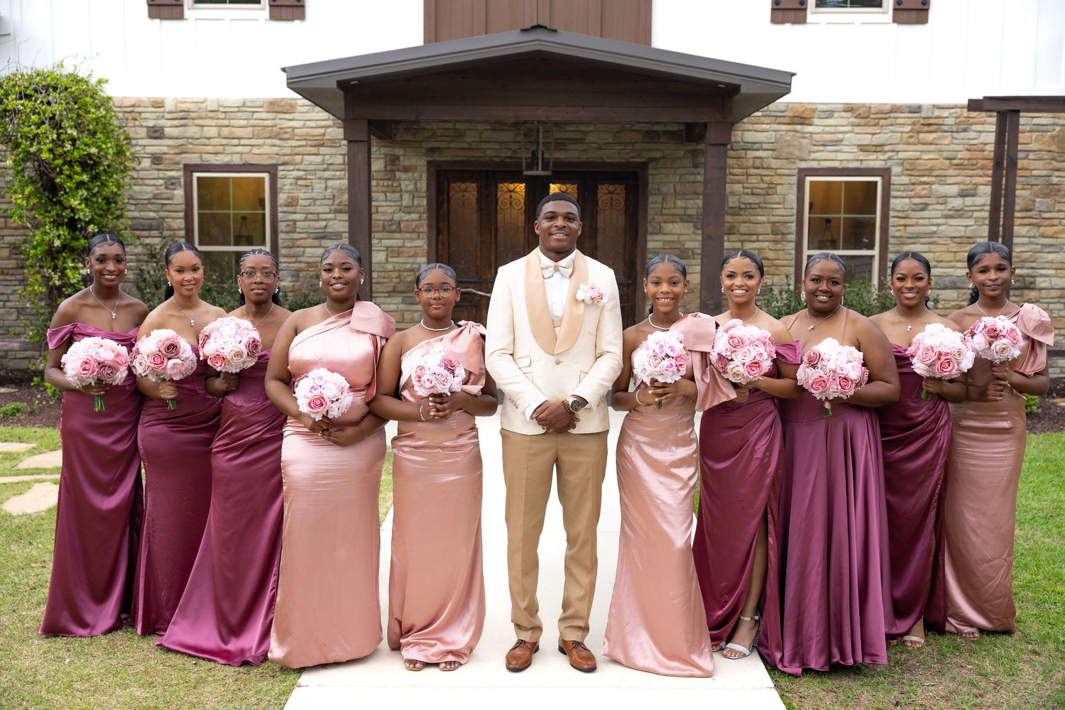 Groom in tan suit poses with bridesmaids in mixed mauve and pink gowns holding pink bouquets outside elegant venue.