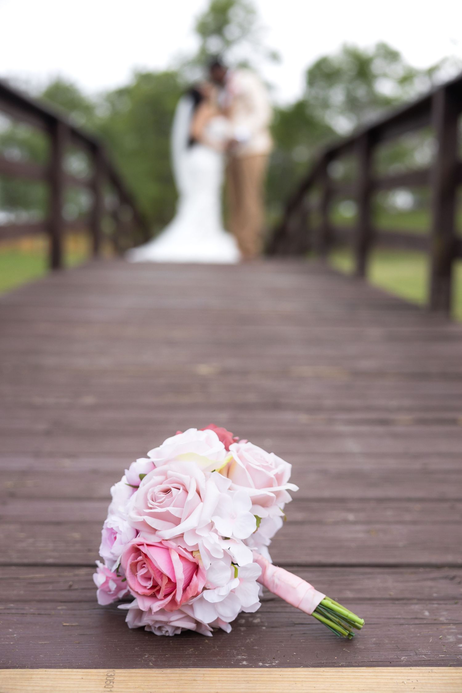 Pink and white rose bridal bouquet on wooden bridge with blurred couple kissing in background.
