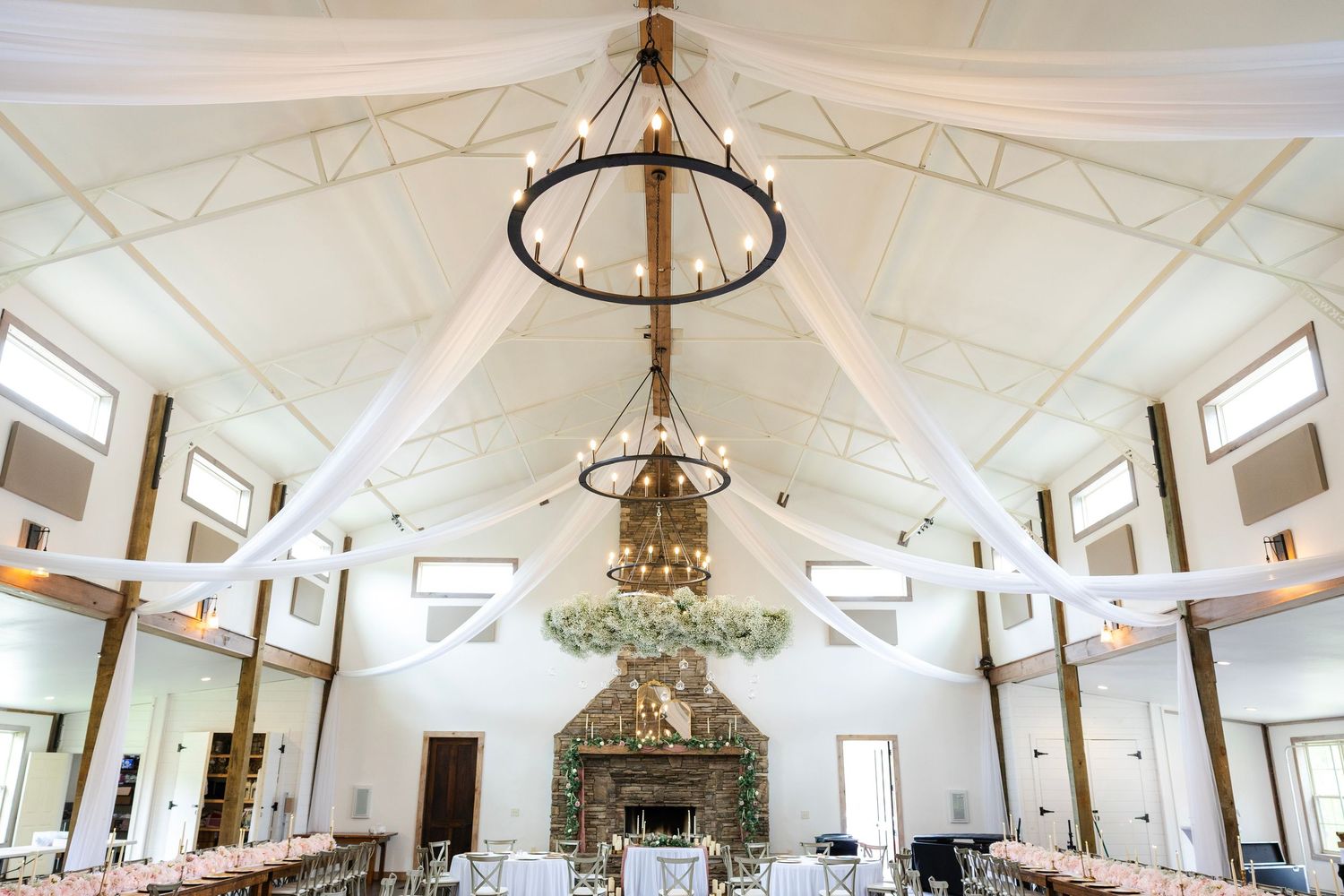 Elegant wedding venue with vaulted white ceiling, chandelier, draped fabric, long reception tables, and a stone fireplace.