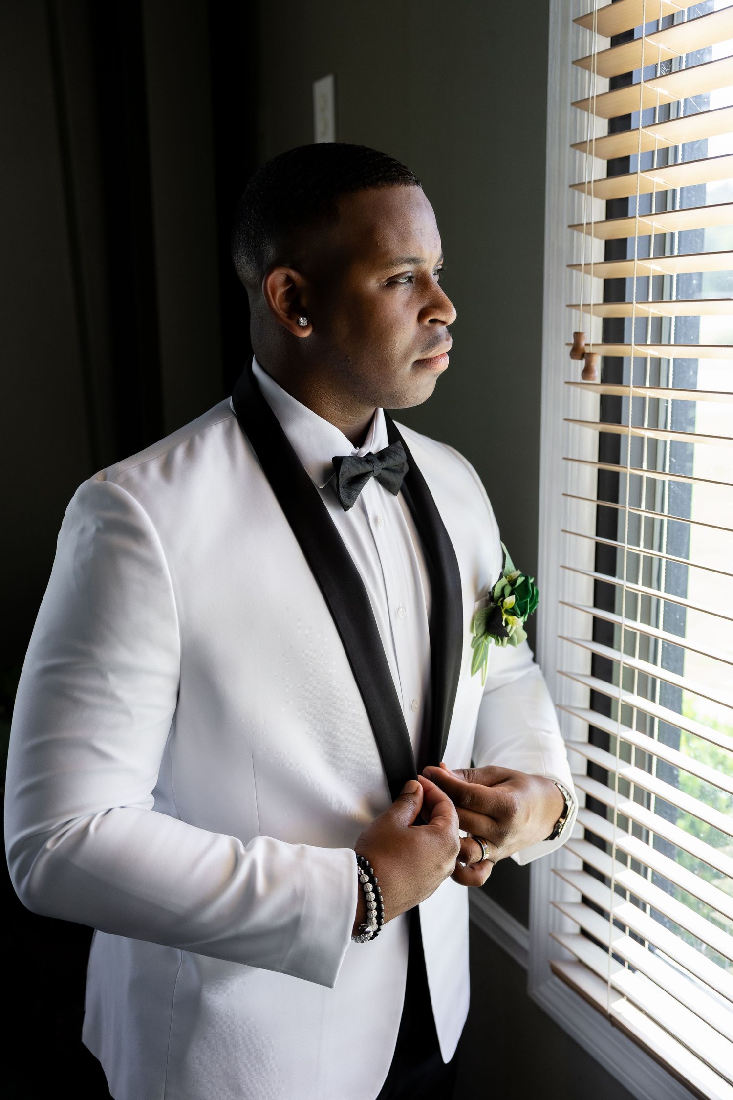 Elegant groom in white tuxedo with black lapels and bow tie adjusting jacket by window with natural light.