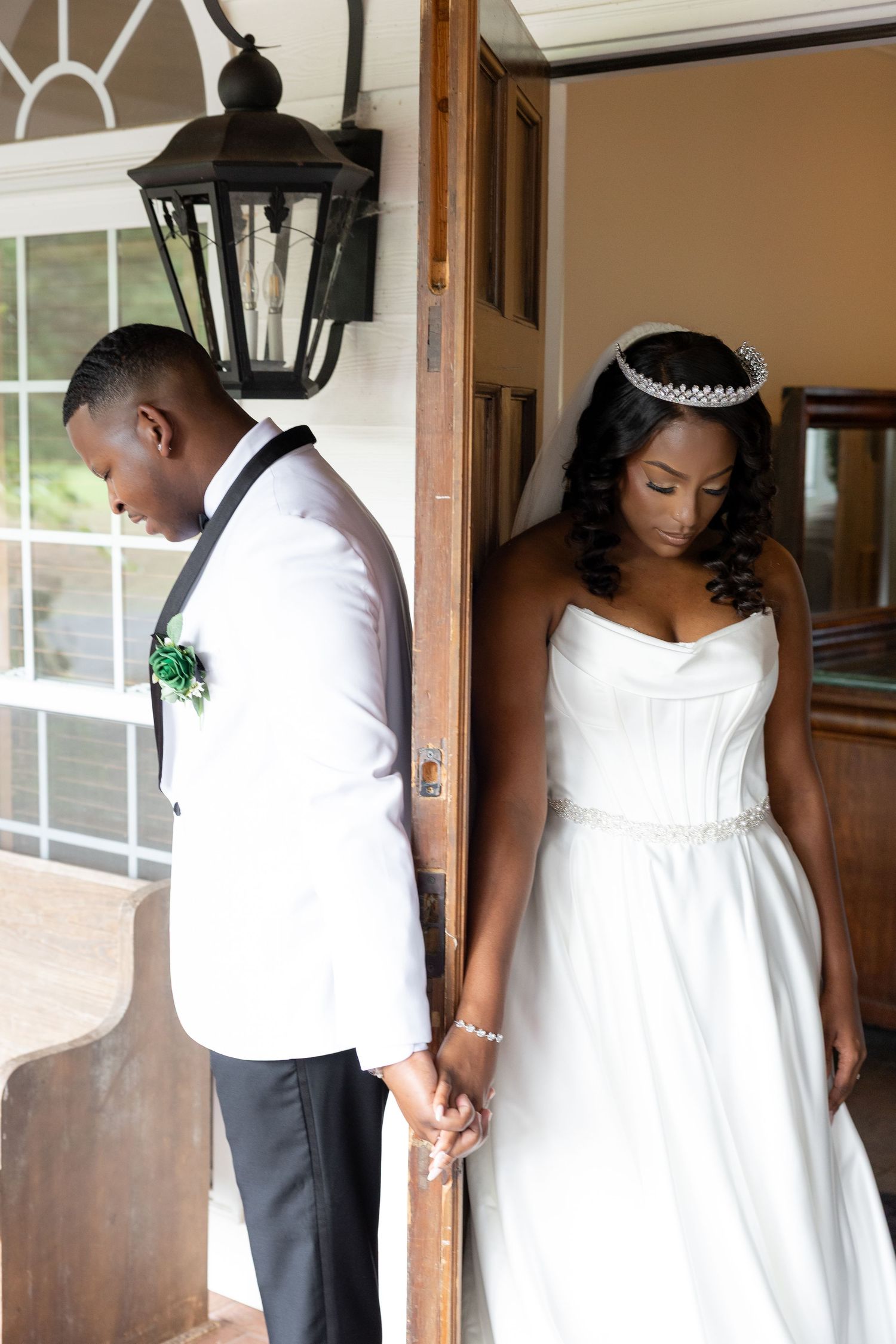 Black bride and groom hold hands through a doorway, she in a white gown and tiara, he in a white tuxedo jacket.