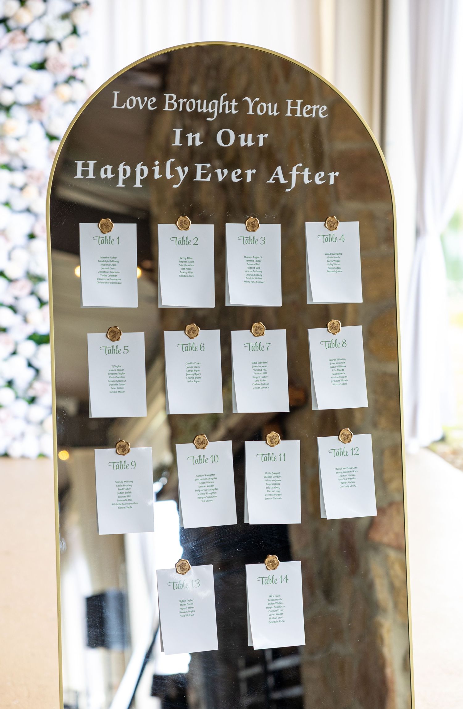 Gold arched mirror wedding seating chart display with white table cards reading Love Brought You Here In Our Happily Ever After.