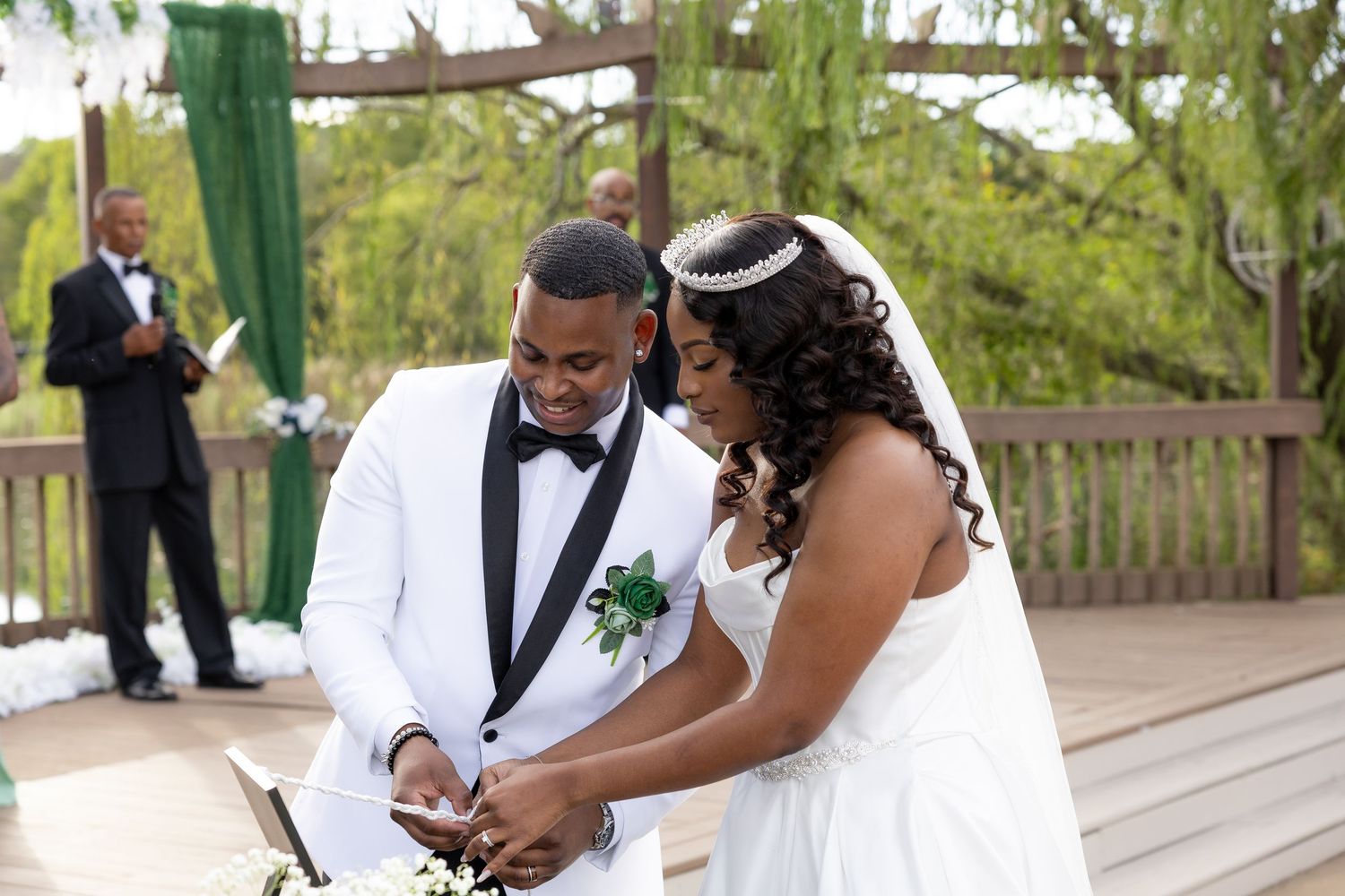 A bride and groom in white wedding attire exchange rings outdoors at a decorated ceremony venue with greenery.