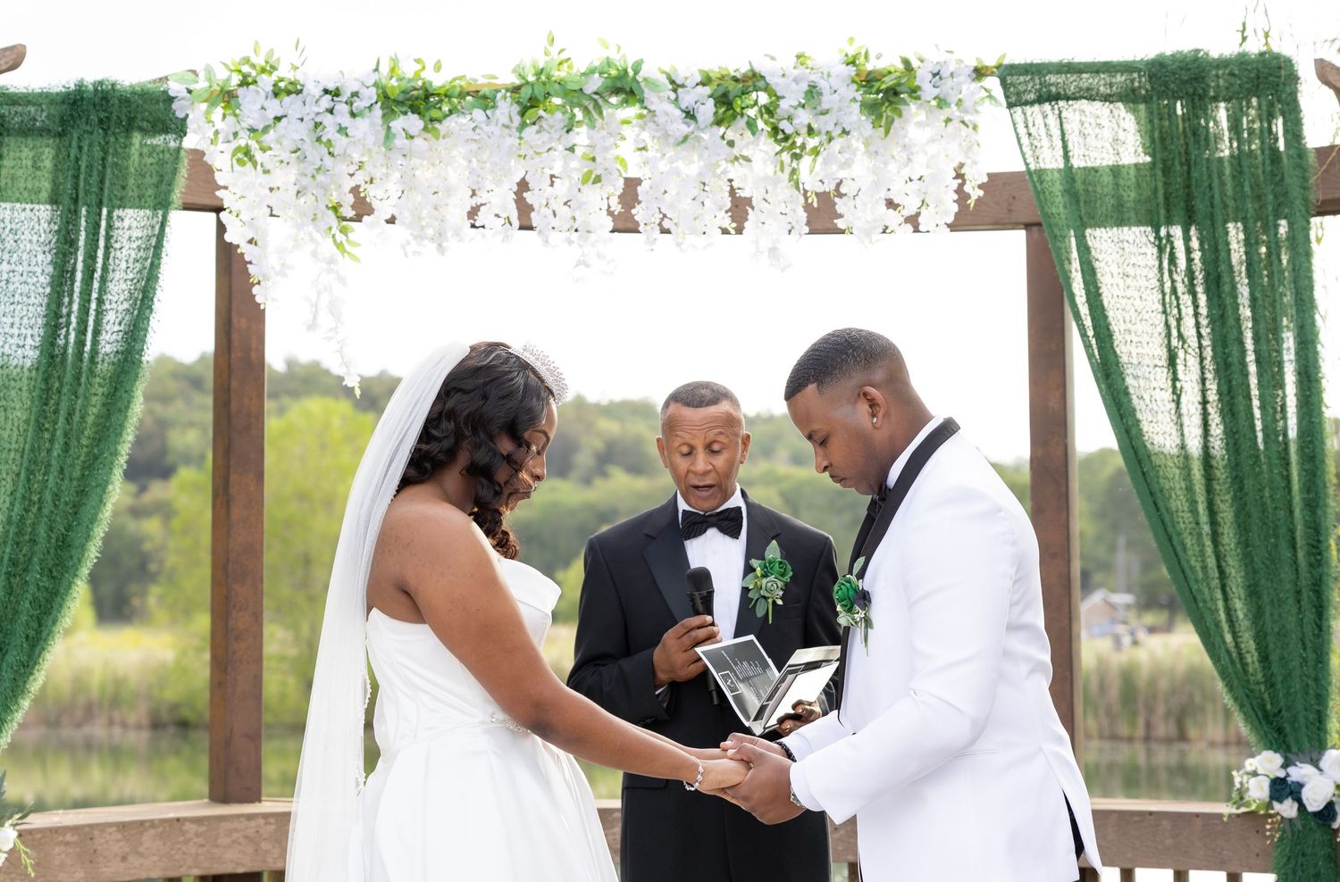 Outdoor wedding ceremony with bride and groom exchanging vows under a floral arch with green drapes.
